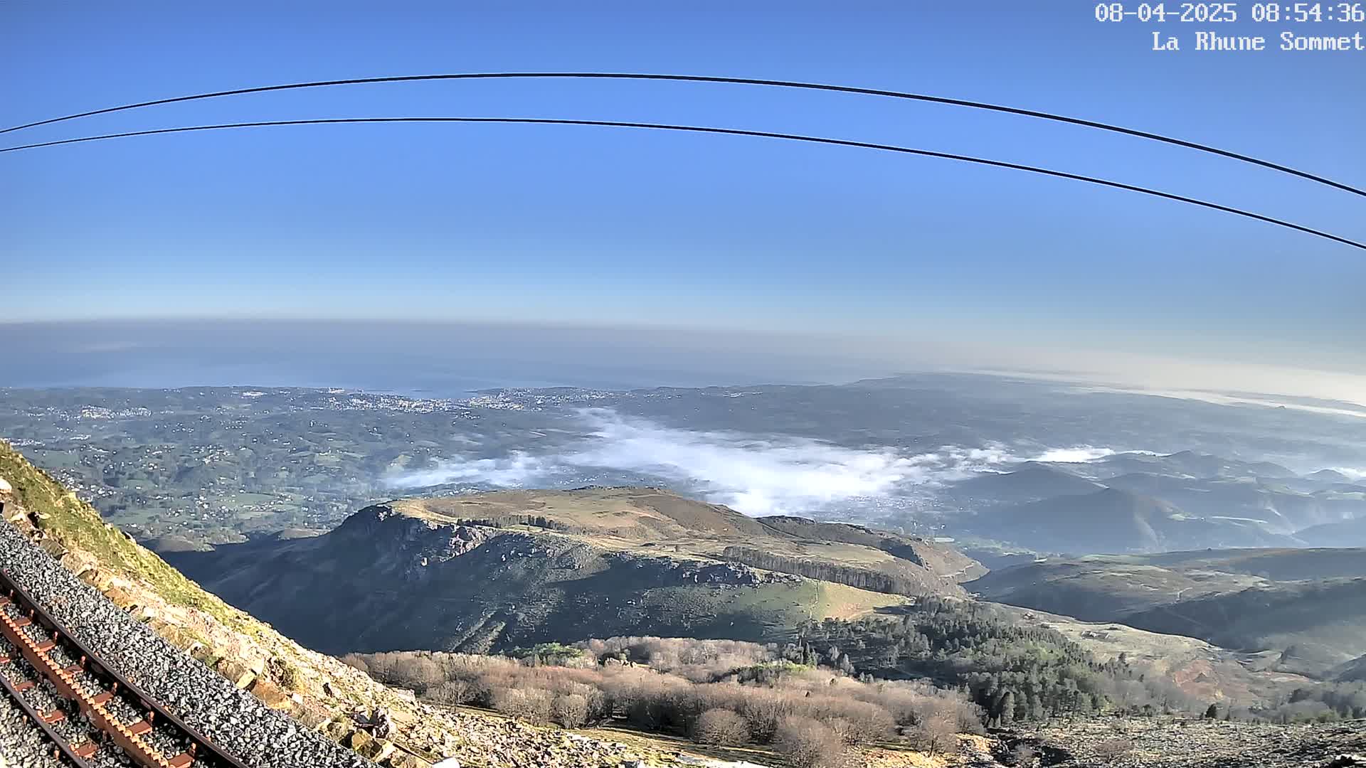 A mountaintop view on a clear day shows a vast expanse of land, a distant ocean, and some low-lying clouds or fog in the valleys below, with train tracks running alongside a rocky slope in the foreground.