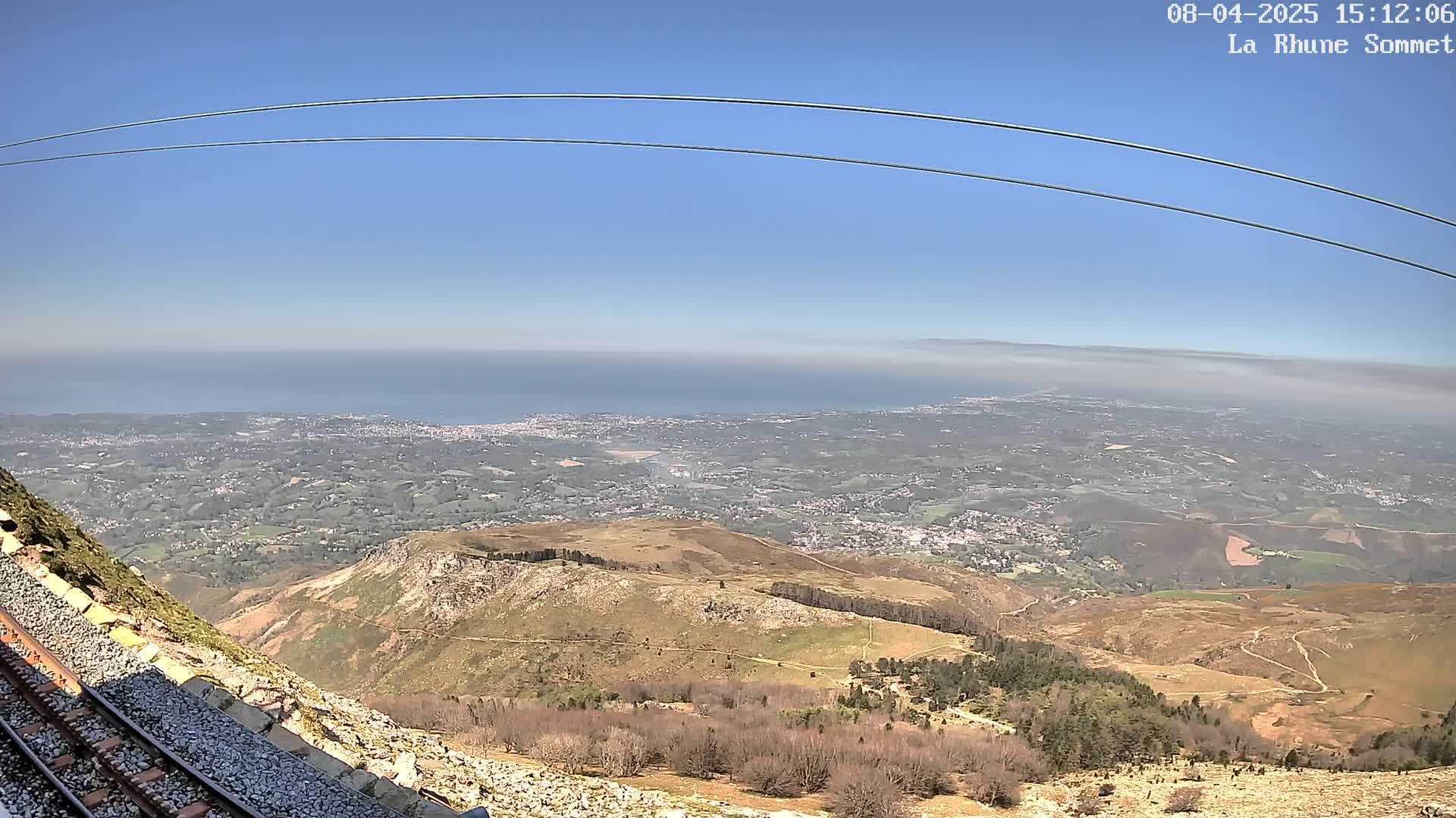 A mountaintop view on a clear day shows a vast expanse of land leading to a hazy ocean, with train tracks and overhead cables visible in the foreground.