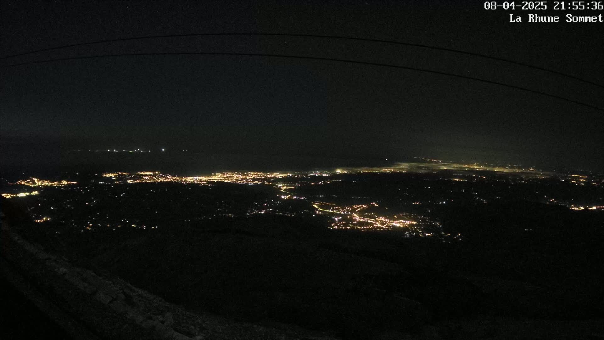 A nighttime view from a high vantage point shows a sprawling city illuminated by scattered lights, extending to a distant coastline under a clear, dark sky.