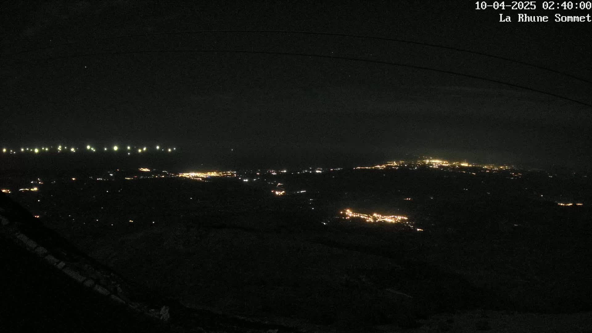 A nighttime, high-angle view shows a sprawling city illuminated by scattered lights, with a line of brighter lights possibly indicating a road or other linear feature in the distance.