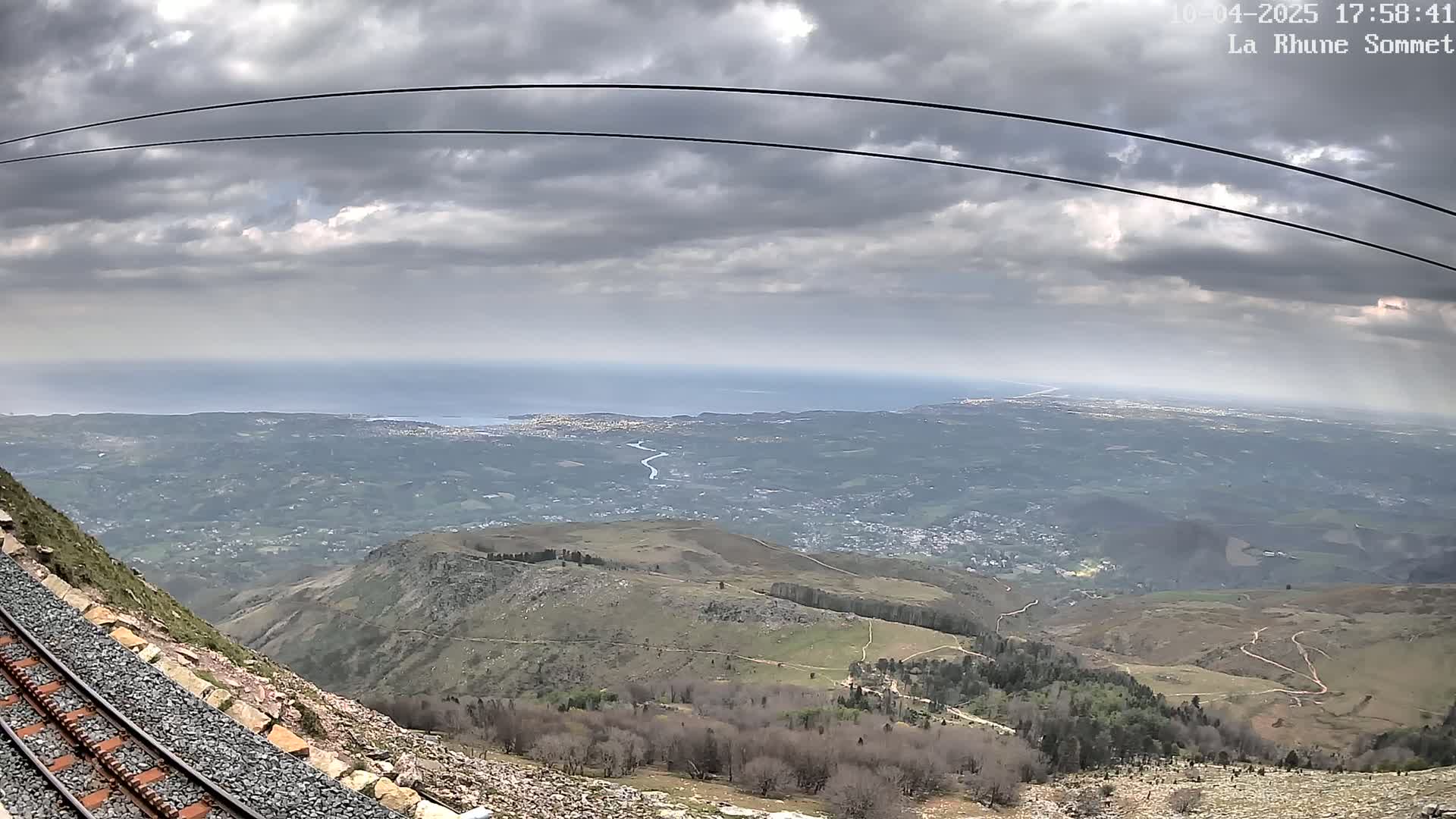A train track runs alongside a mountain overlooking a vast expanse of land, a distant ocean, and a cloudy sky.