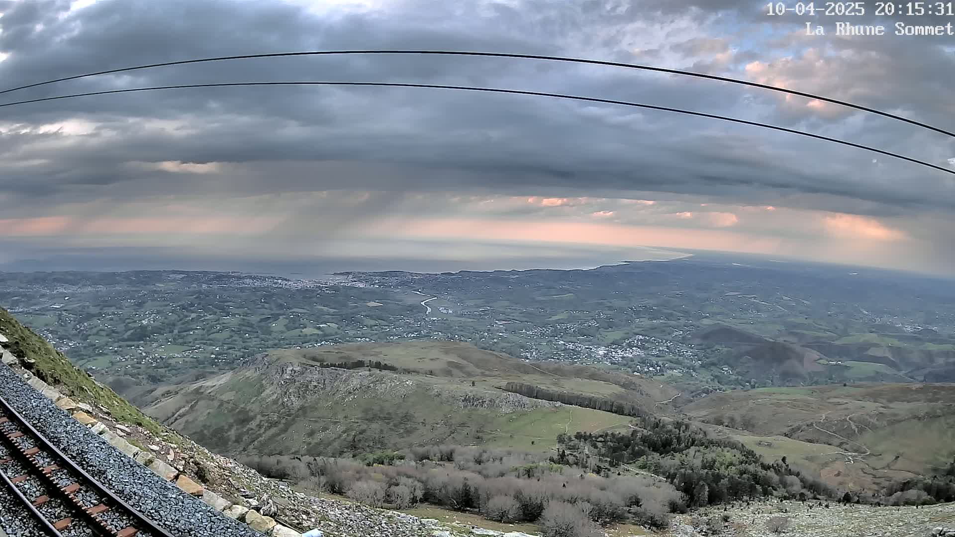 A mountaintop view shows a railway track leading down a steep hillside toward a vast, sprawling coastal town under a cloudy, overcast sky with hints of pink at the horizon.