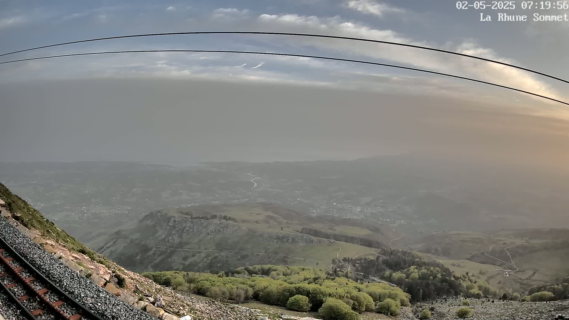 A hazy mountain vista is seen from a high vantage point, with a railway track and cable lines visible in the foreground under a partly cloudy sky.