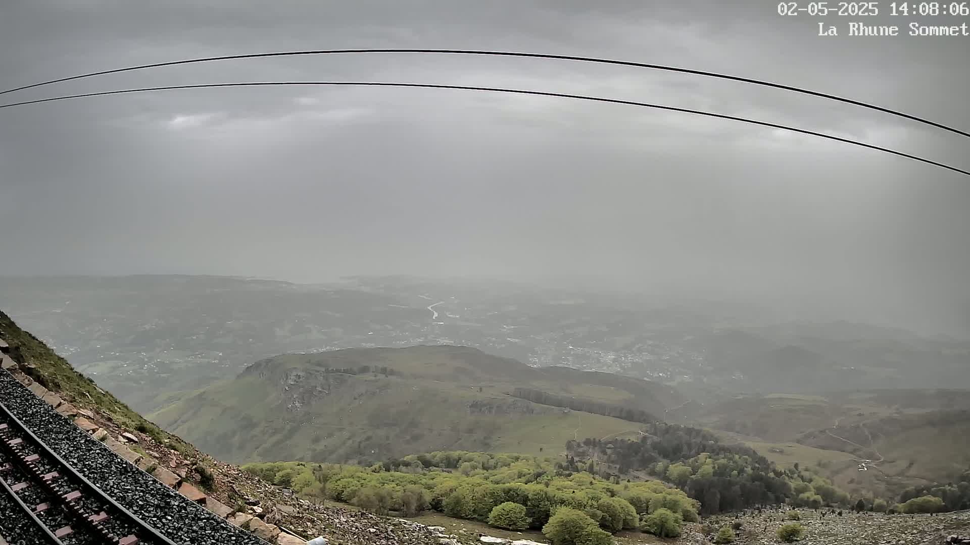 A hazy view of a valley and distant town is seen from a mountainside railway track under an overcast sky.