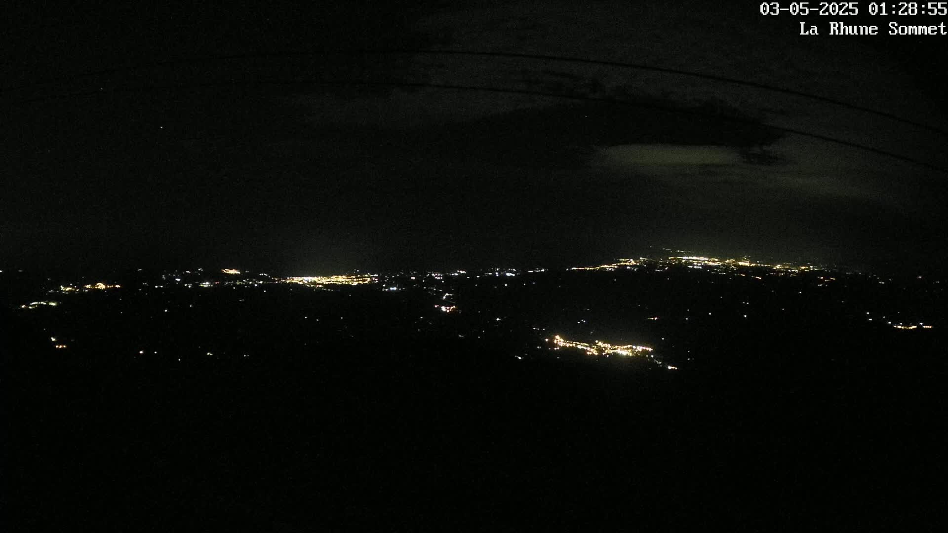 A nighttime, long-range view of a distant city’s lights spread across a dark landscape under a cloudy sky.