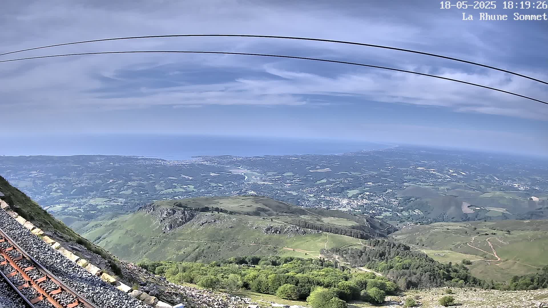 A mountaintop view reveals a vast expanse of green hills and valleys extending to a distant coastline under a partly cloudy sky, with train tracks visible in the foreground.