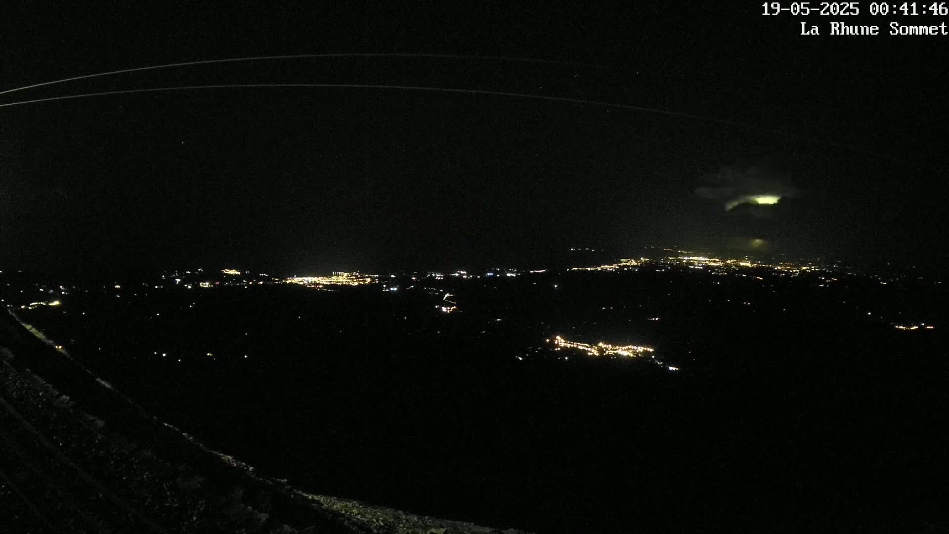 A nighttime view from a high vantage point shows a distant city illuminated below, with a faint, glowing cloud formation in the sky.