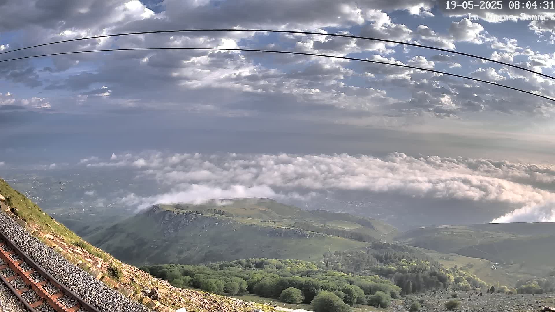 A mountaintop view shows a railway track beside a rocky hillside, overlooking a valley covered in low clouds under a partly cloudy sky.
