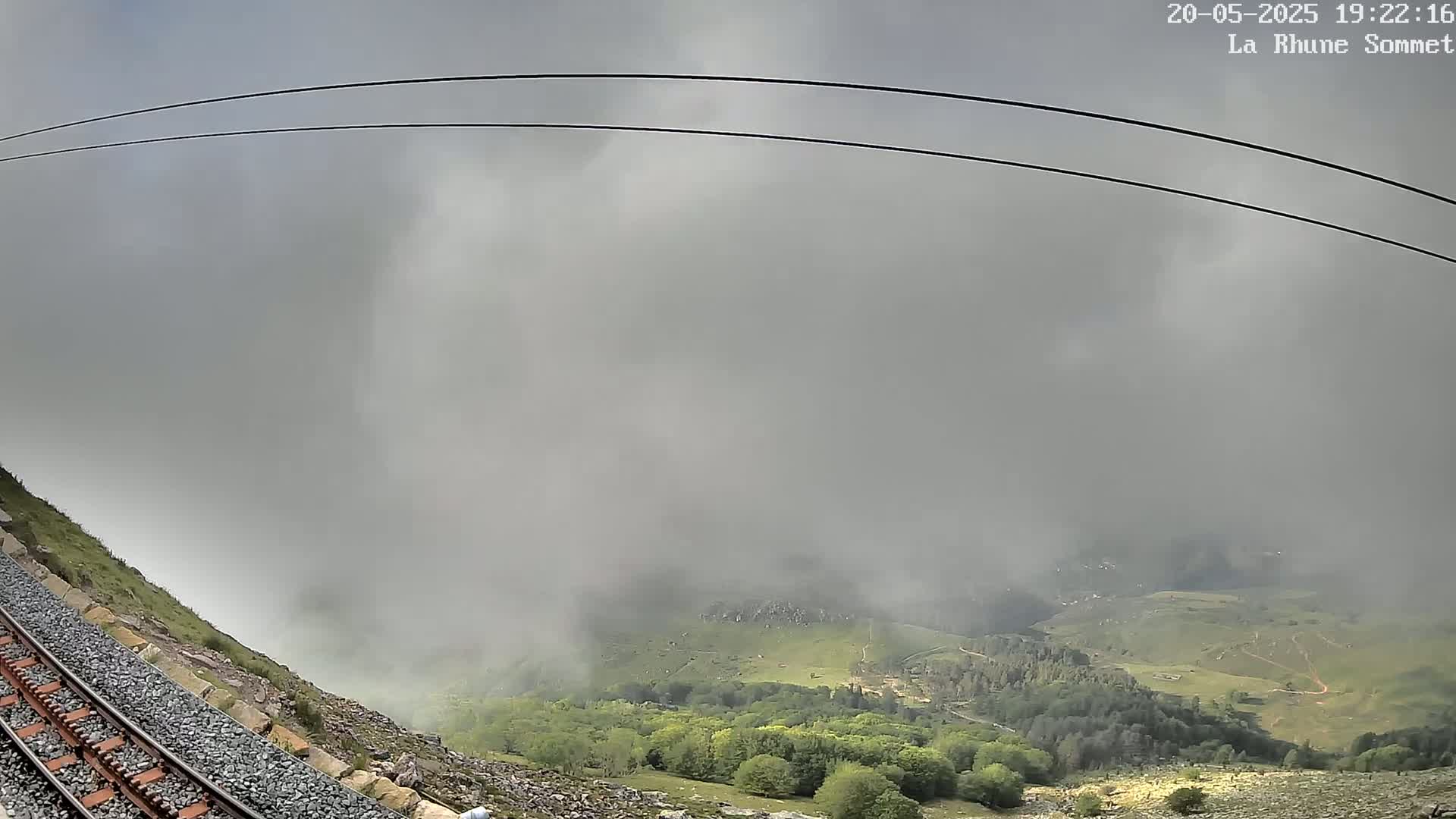 A railway track runs alongside a rocky hillside, descending into a foggy valley with low-lying vegetation.