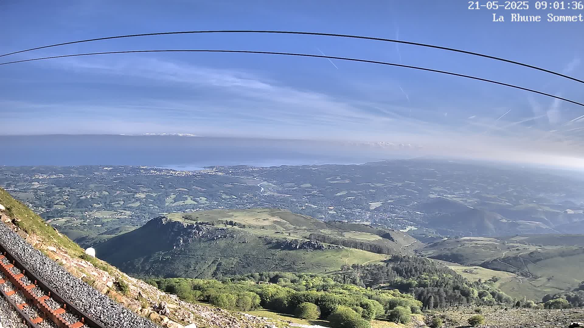 A mountaintop view on a clear day shows a vast expanse of rolling hills and valleys leading to a distant body of water with mountains on the horizon, all viewed alongside a cable car track.