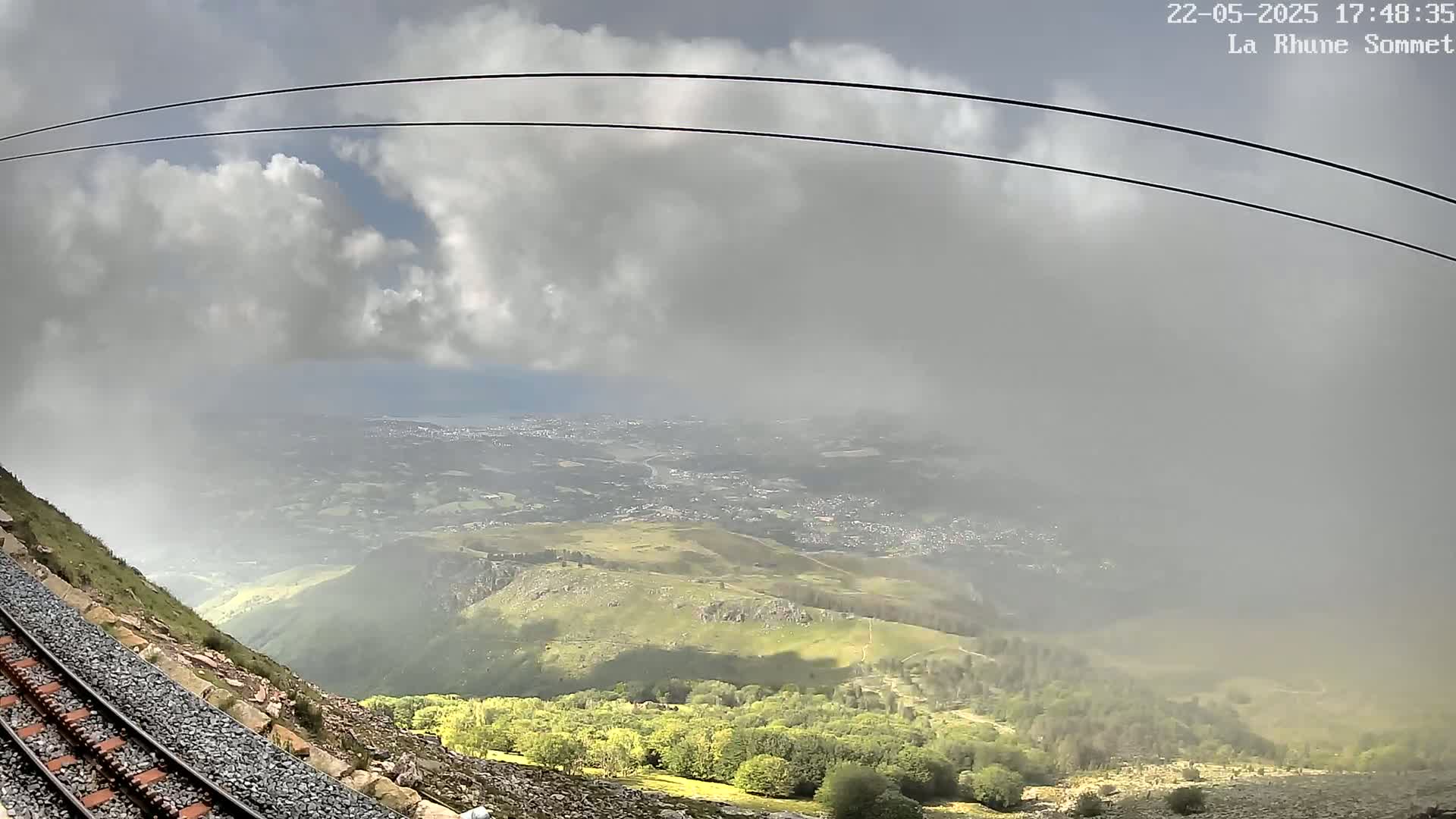 A mountain railway track descends a steep hillside towards a valley partially obscured by low clouds and mist, with a distant town visible beneath a partly cloudy sky.