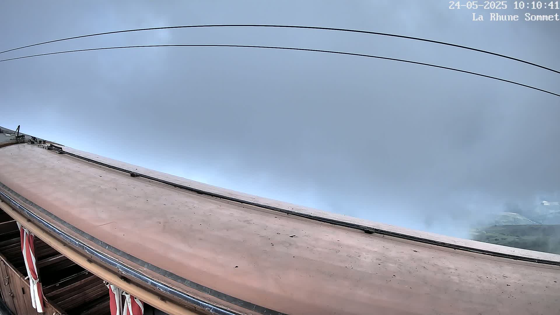 A tan-colored train car's roof and upper sides are visible, with overcast skies and distant, hazy greenery in the background.