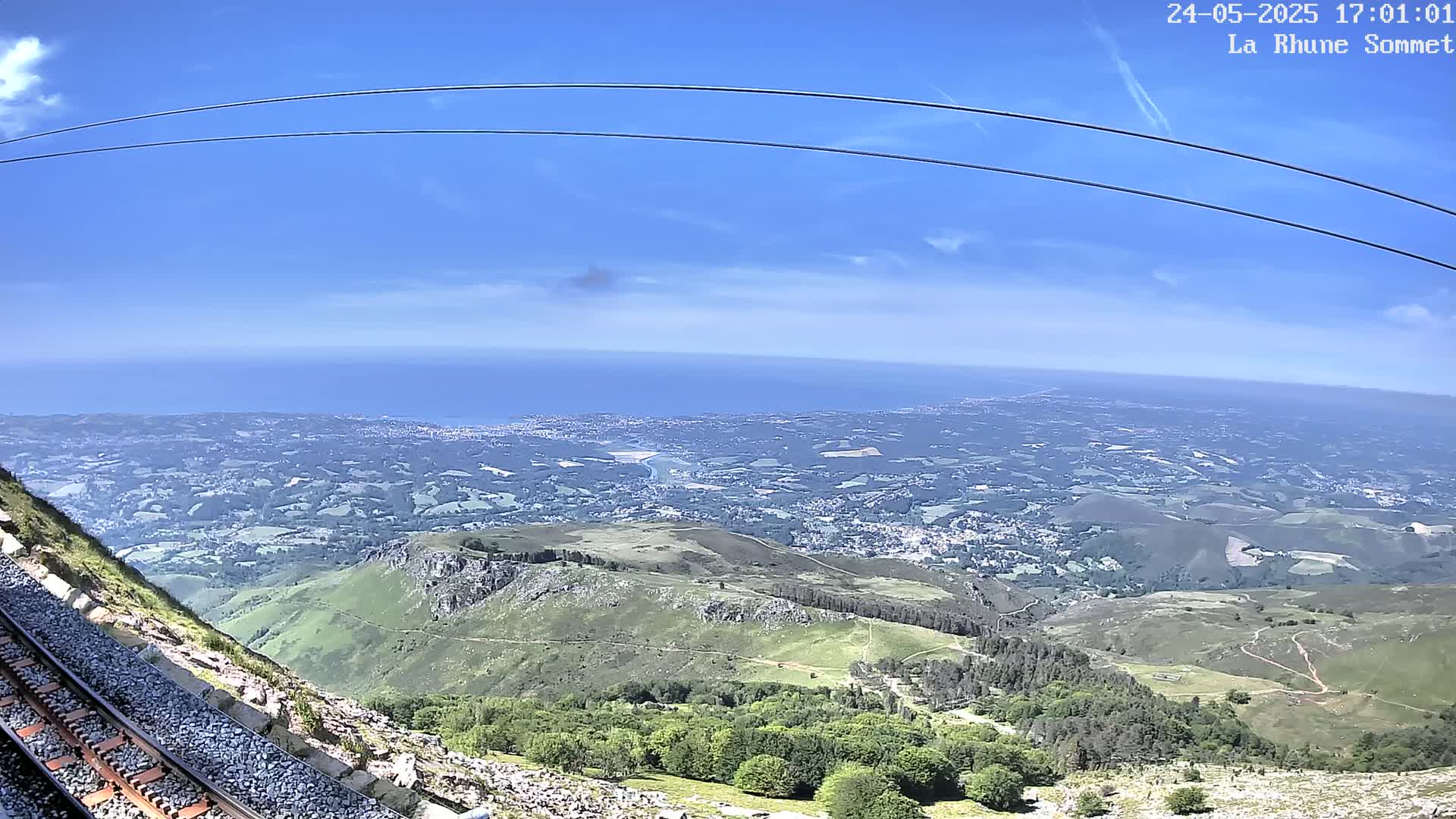 A mountaintop view on a clear, sunny day shows a vast expanse of land stretching to a distant ocean, with train tracks running alongside a rocky hillside.