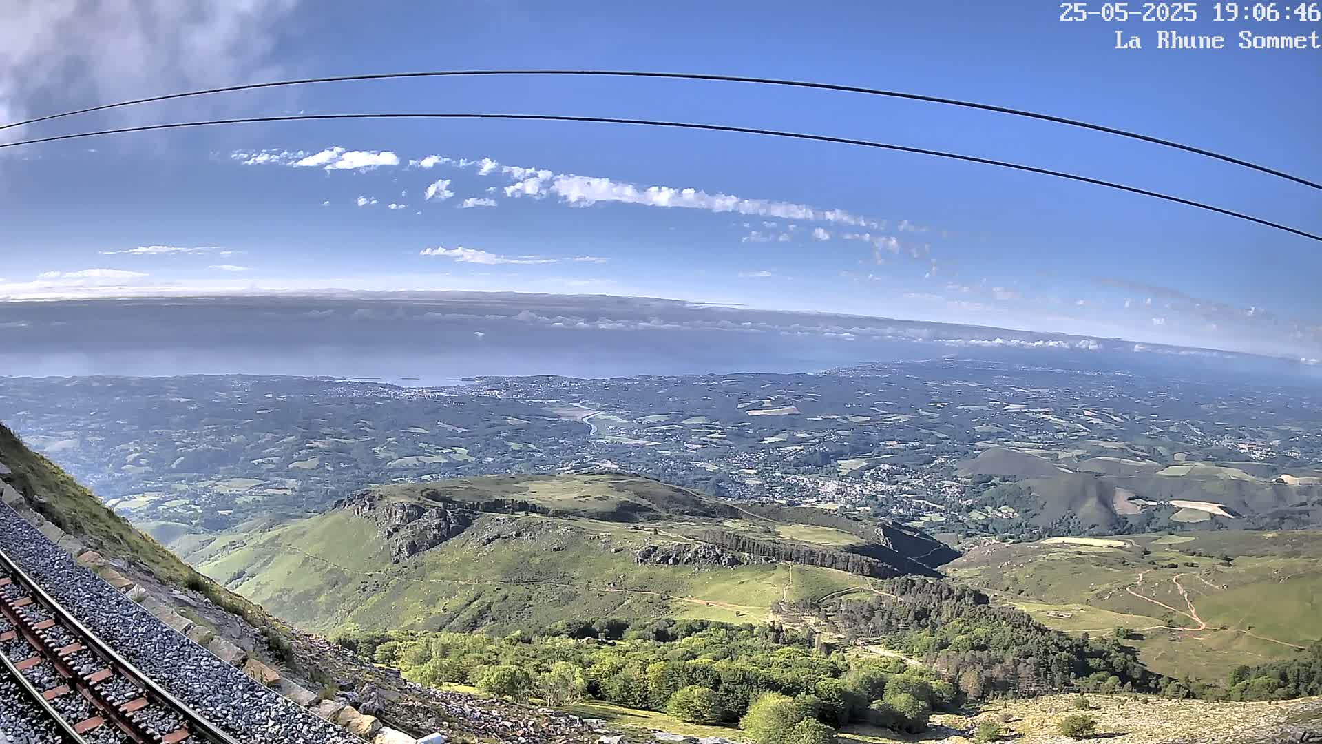A mountaintop view on a partly cloudy day shows a train track leading down a hillside toward a vast expanse of green valleys and a distant ocean.