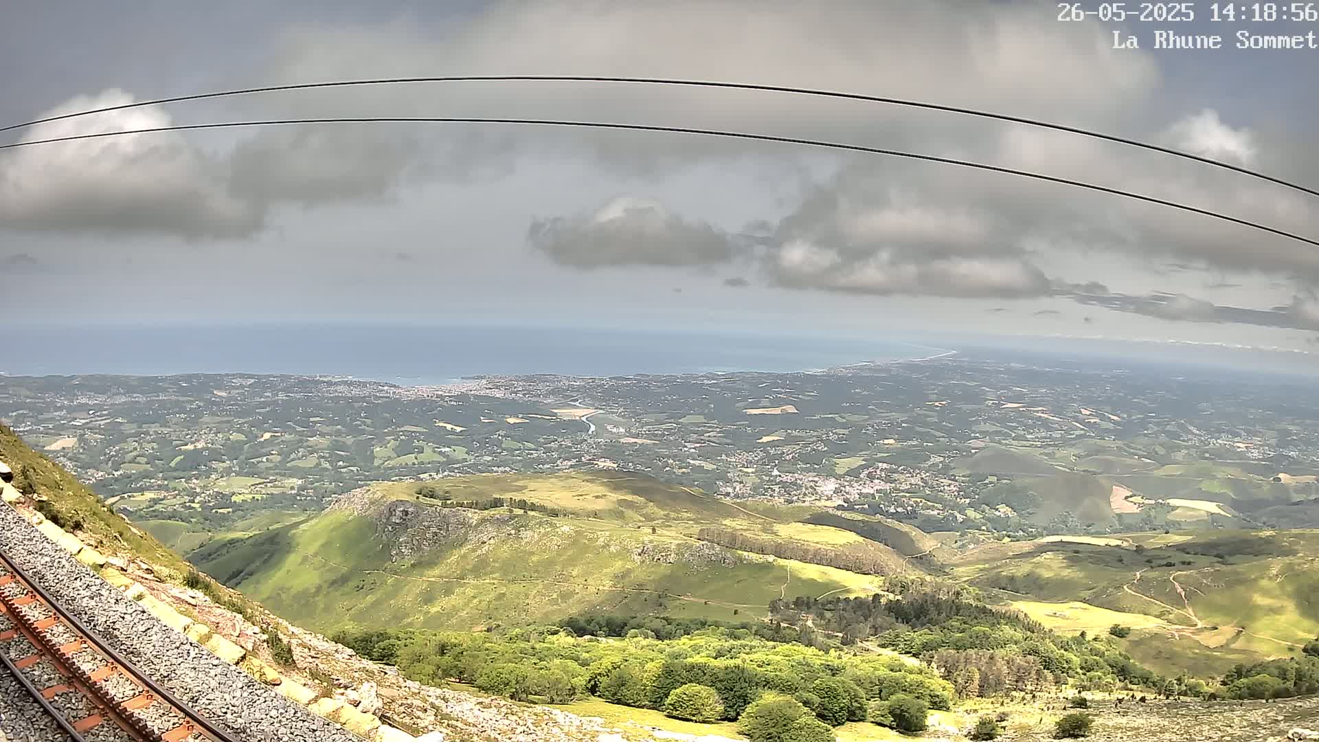A mountaintop view shows a vast expanse of green hills and valleys extending to a distant coastline under a partly cloudy sky.