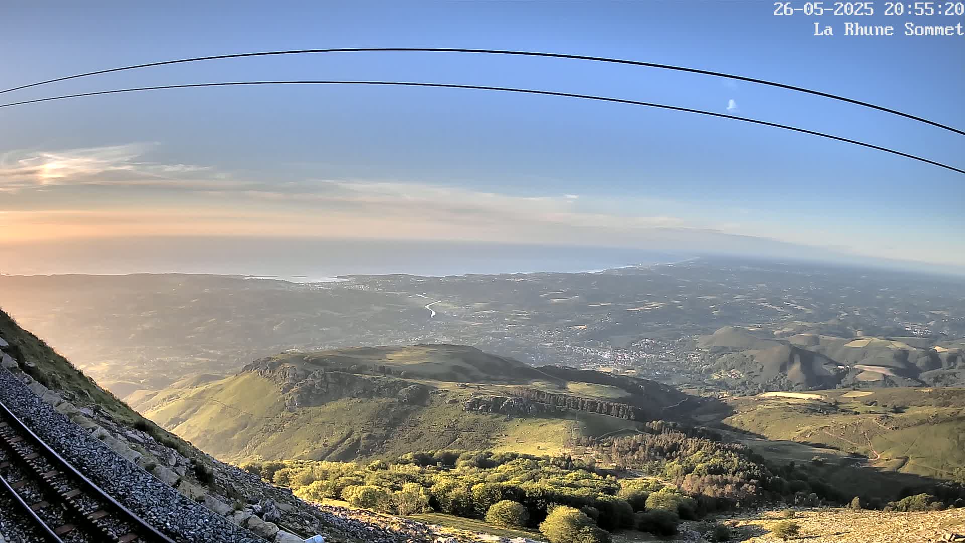 A mountaintop view at sunset shows a vast expanse of land extending to a calm ocean under a clear, light blue sky, with train tracks and cable wires visible in the foreground.