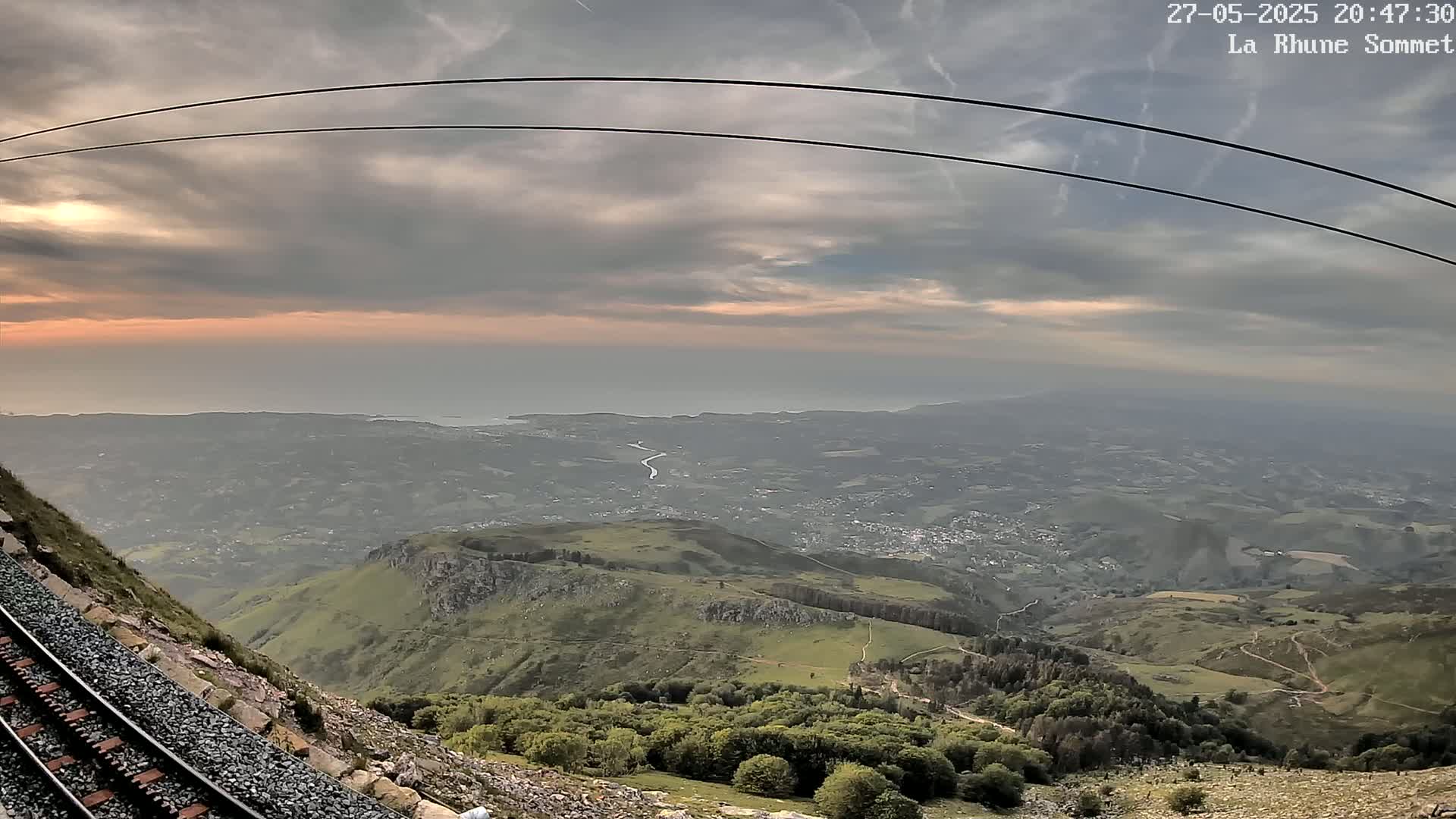 A mountaintop railway track overlooks a vast, hilly landscape extending to a calm ocean under a mostly cloudy sunset sky.