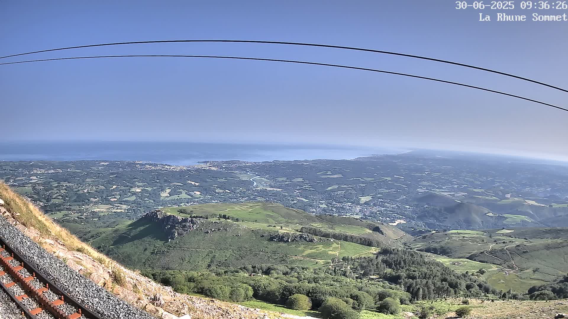 A mountainside railway track overlooks a vast expanse of green hills and valleys leading to a distant ocean under a clear blue sky.