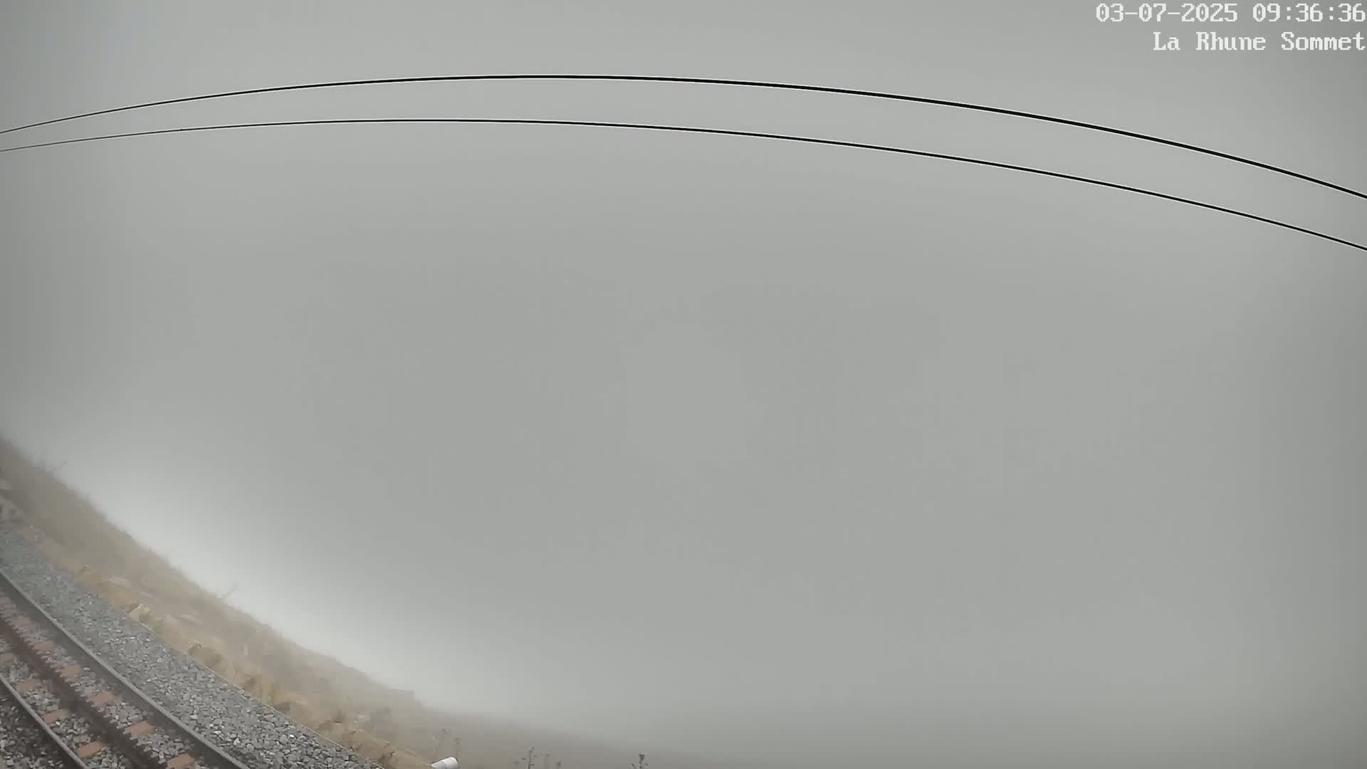 A view from a train shows a thick fog obscuring the landscape, with train tracks visible in the foreground and power lines overhead.