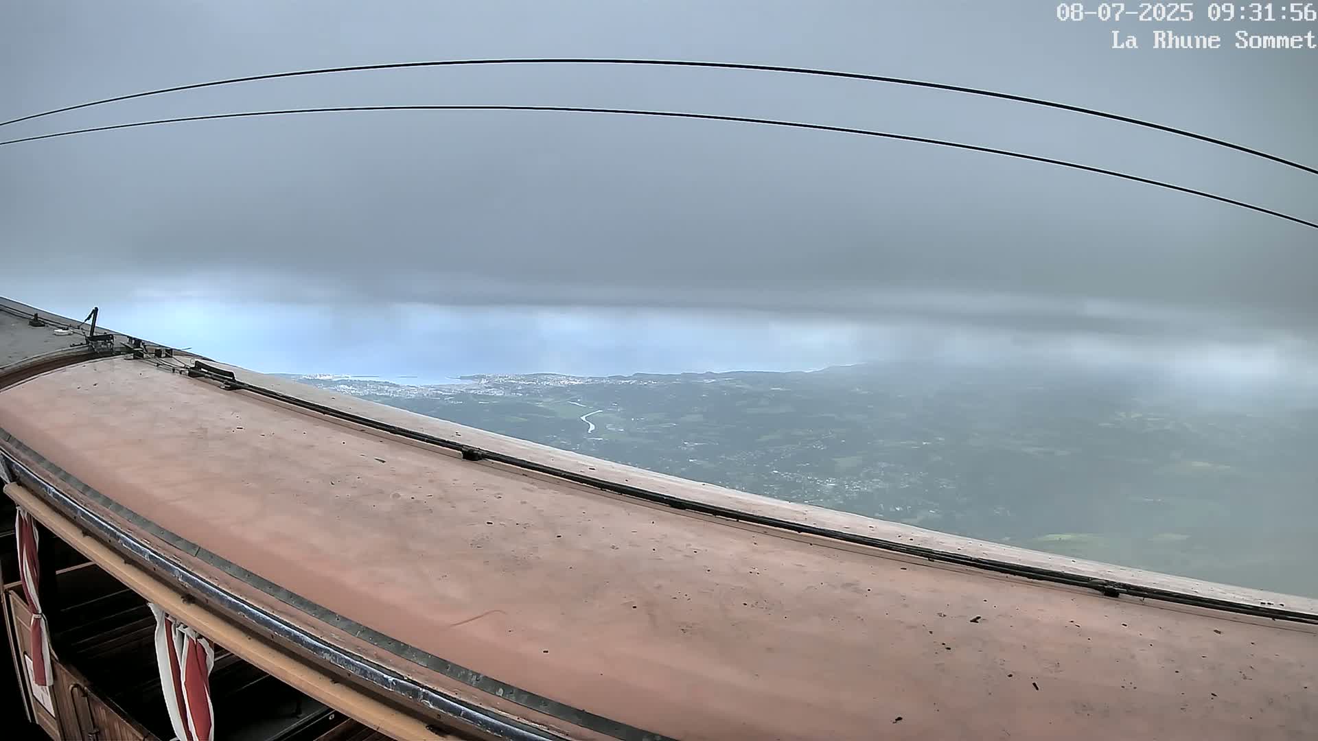 A cable car's roof and a view of overcast skies and distant land below.