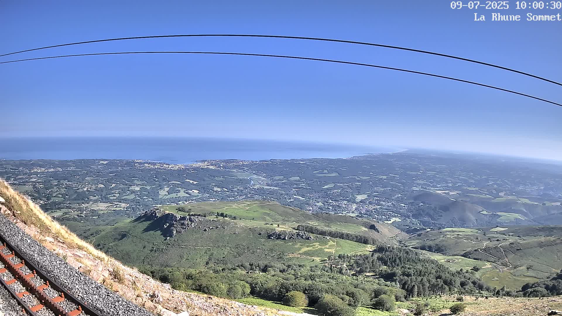 A mountaintop view on a clear day shows a vast expanse of land stretching to a distant ocean, with train tracks visible in the foreground.