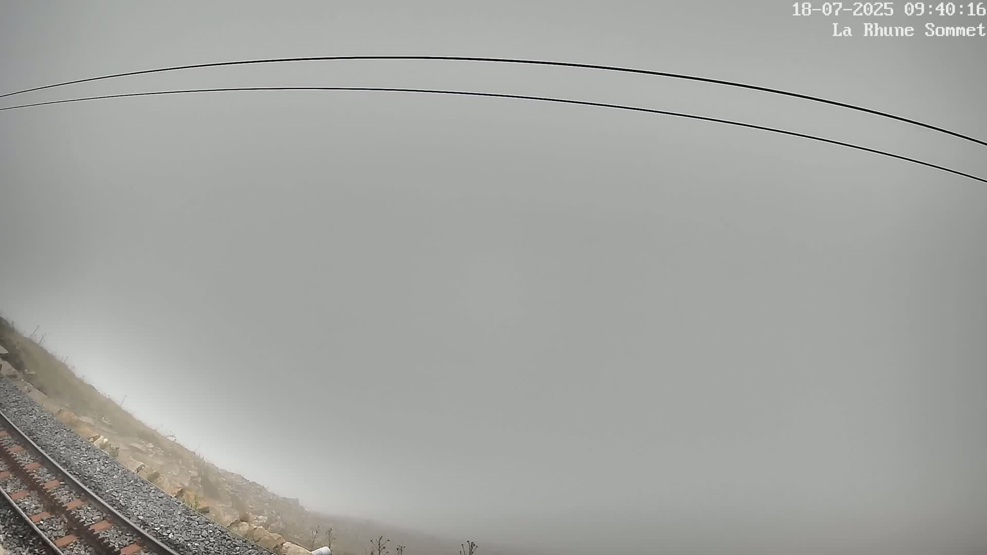 A railway track runs alongside a rocky hillside shrouded in thick fog, with power lines arching overhead.