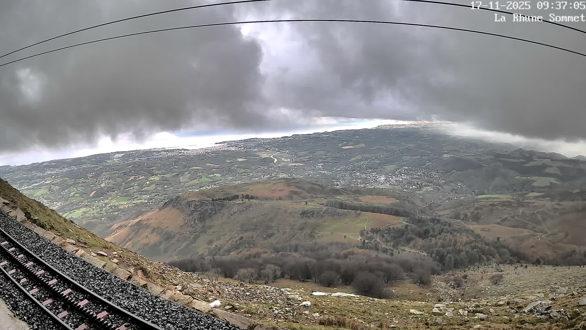 A wide, high-angle view captures rack railway tracks descending a rocky mountain slope, overlooking a sprawling panorama of green valleys, distant towns, and a coastline, all under a heavily overcast and gloomy sky.