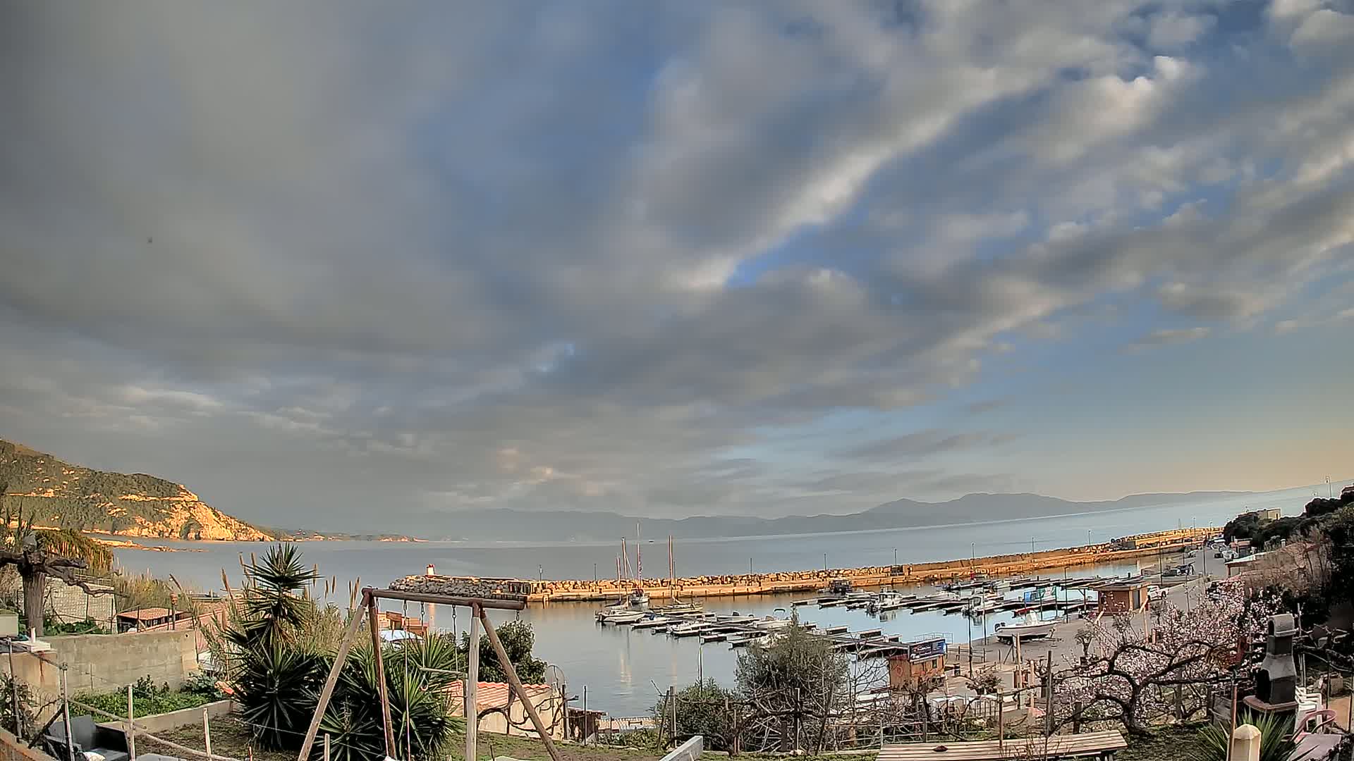A mostly cloudy sky overlooks a calm bay filled with small boats, a stone breakwater, and a small village nestled next to a hillside.