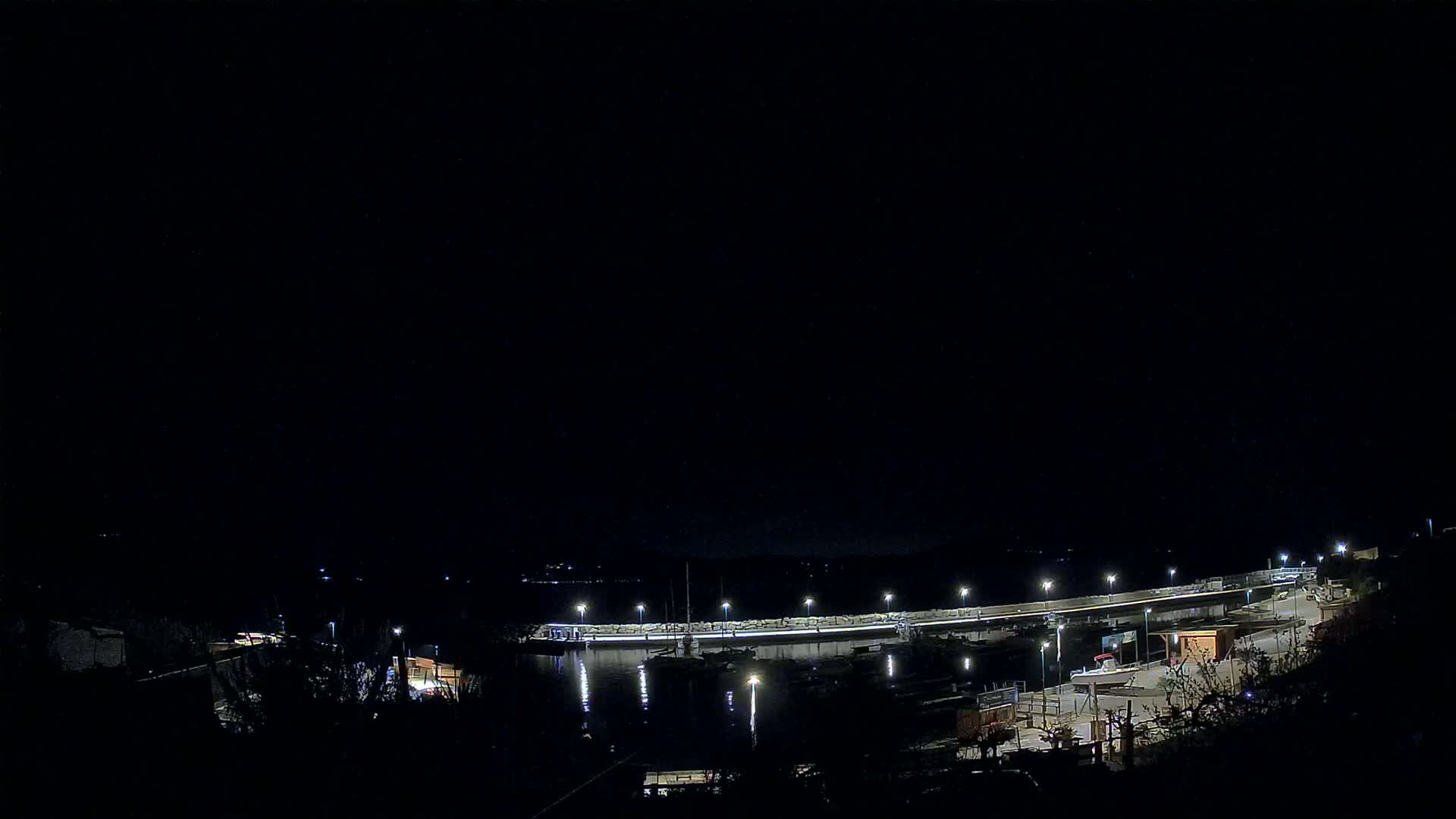 A nighttime view of a harbor with boats docked along a lit seawall under a clear, dark sky.