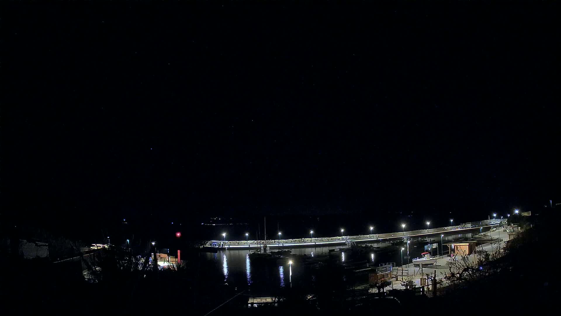 A nighttime view of a harbor with boats docked along a lit seawall under a clear, starry sky.
