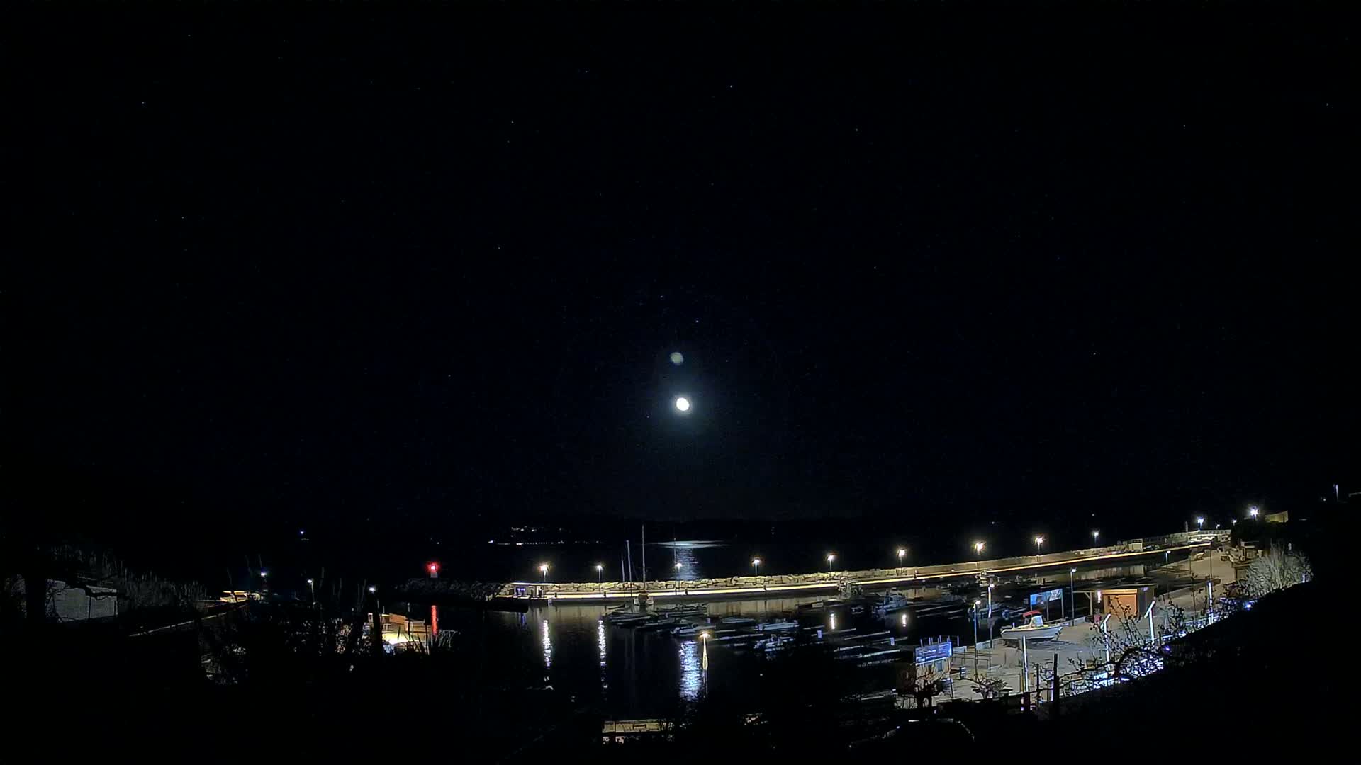 A moonlit night scene shows a harbor filled with boats, illuminated by lights along a curved wall, under a clear, starry sky.