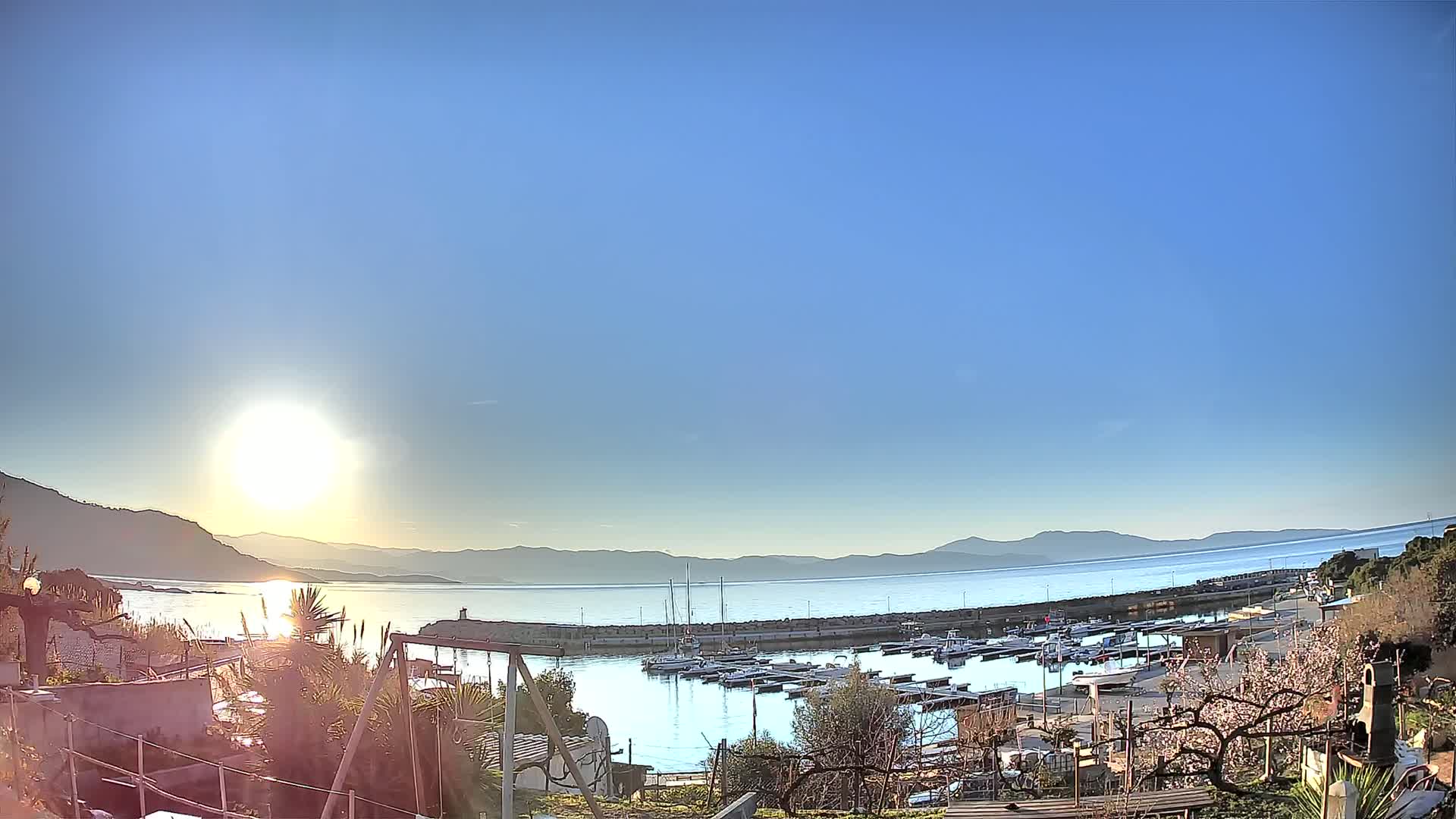 A calm, sunny day reveals a marina filled with boats, nestled between a hillside and a tranquil body of water stretching towards distant mountains.