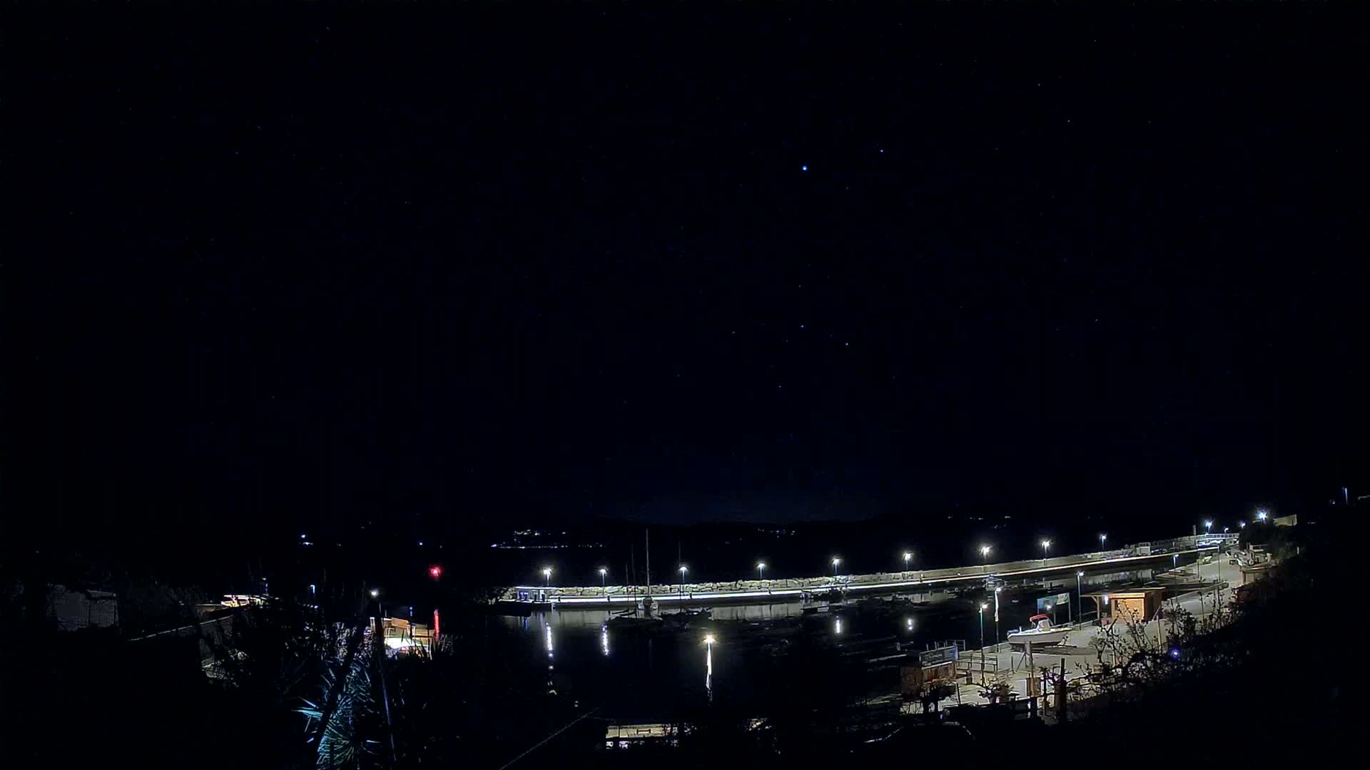 A nighttime view of a marina with boats docked along a lit seawall under a clear, starry sky.