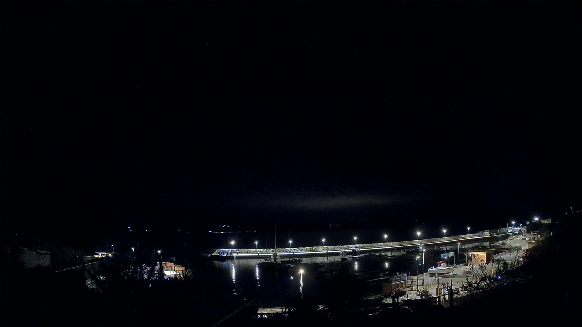 A nighttime, low-angle view of a marina with boats docked along a lit seawall under a dark, clear sky.