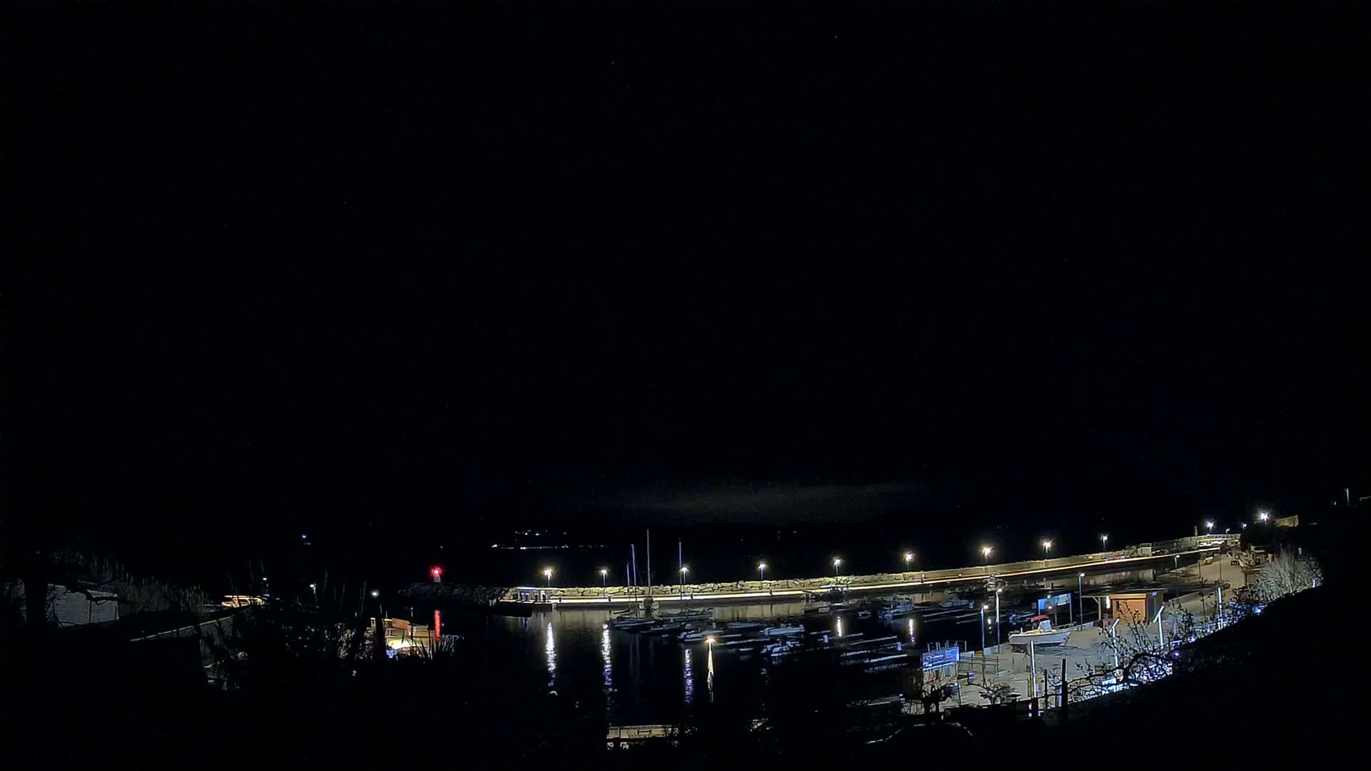 A nighttime view of a marina with numerous boats docked along a lit seawall under a clear, dark sky.