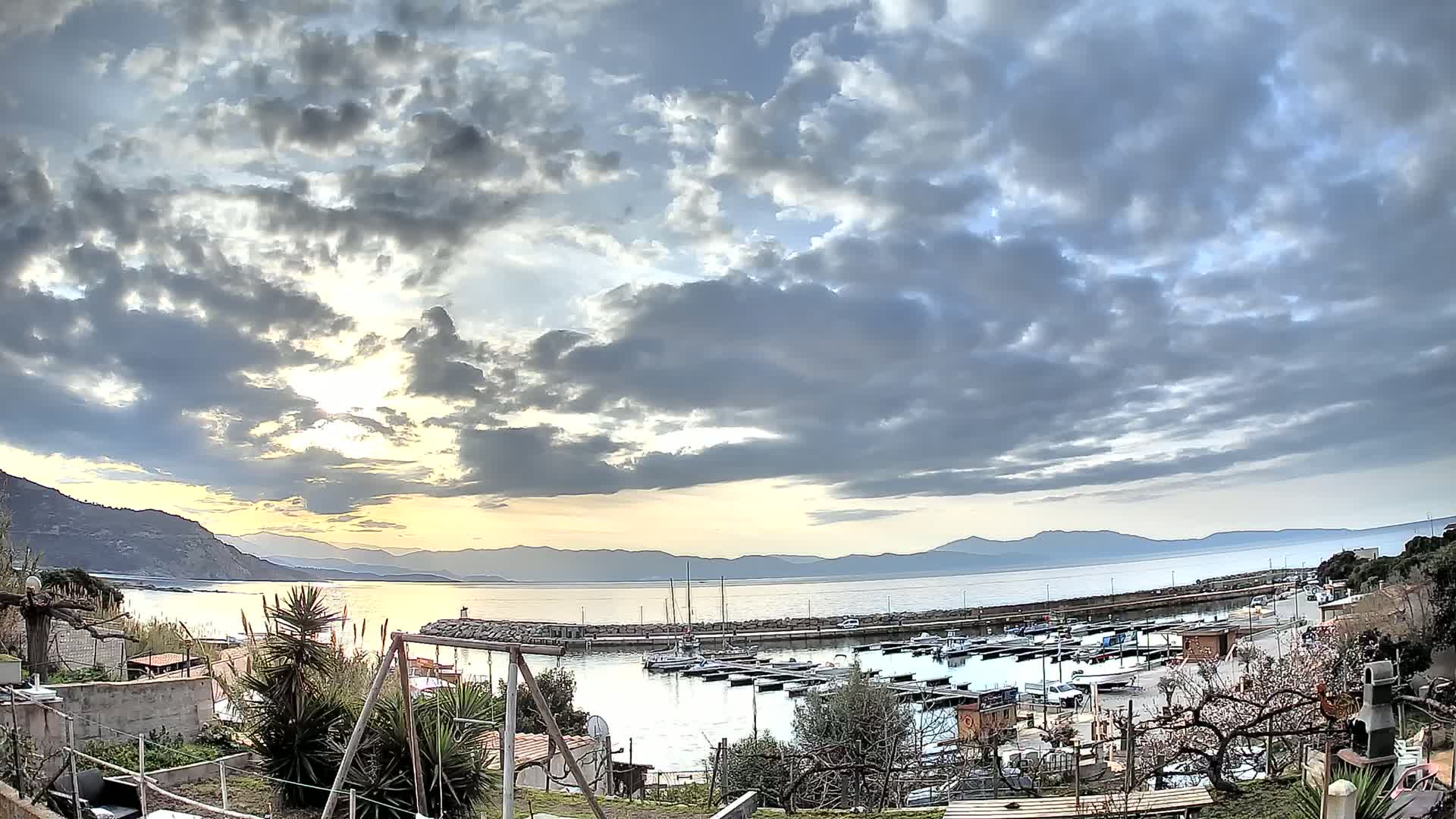 A mostly cloudy sunset over a calm bay with a small marina filled with boats, viewed from a hillside garden.