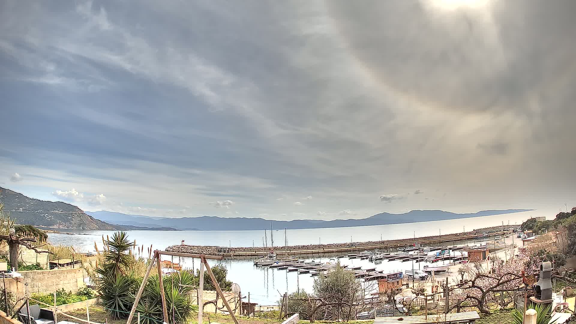 A small harbor filled with boats is seen from a hillside under a mostly cloudy sky.