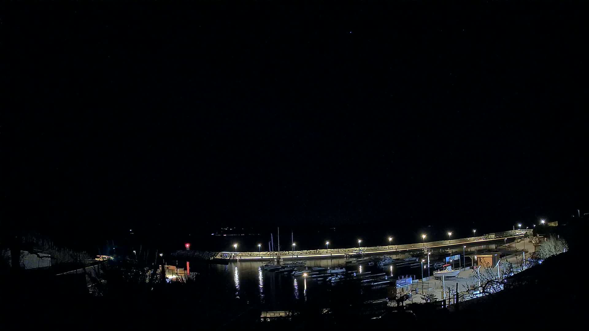 A nighttime view of a harbor filled with boats, illuminated by lights along a seawall under a clear, dark sky.