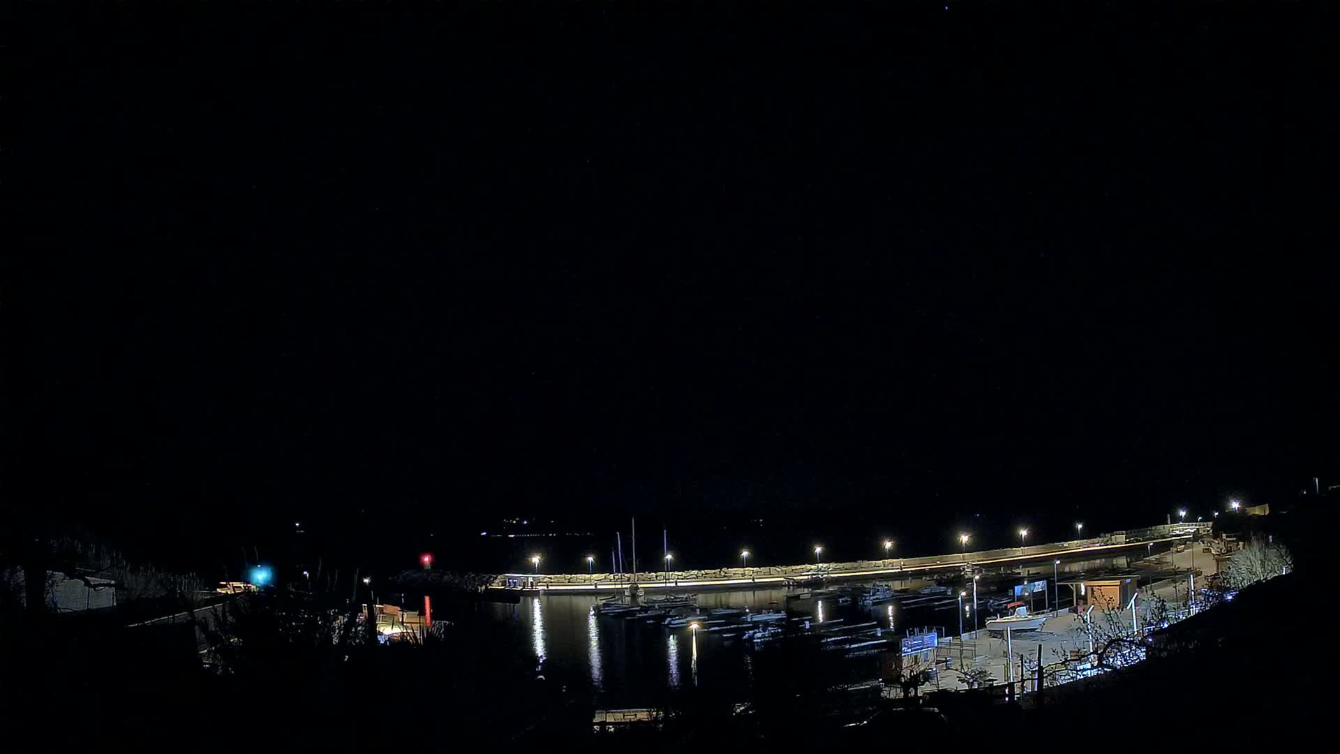 A nighttime view of a harbor filled with boats, illuminated by lights along a curved wall under a clear, dark sky.