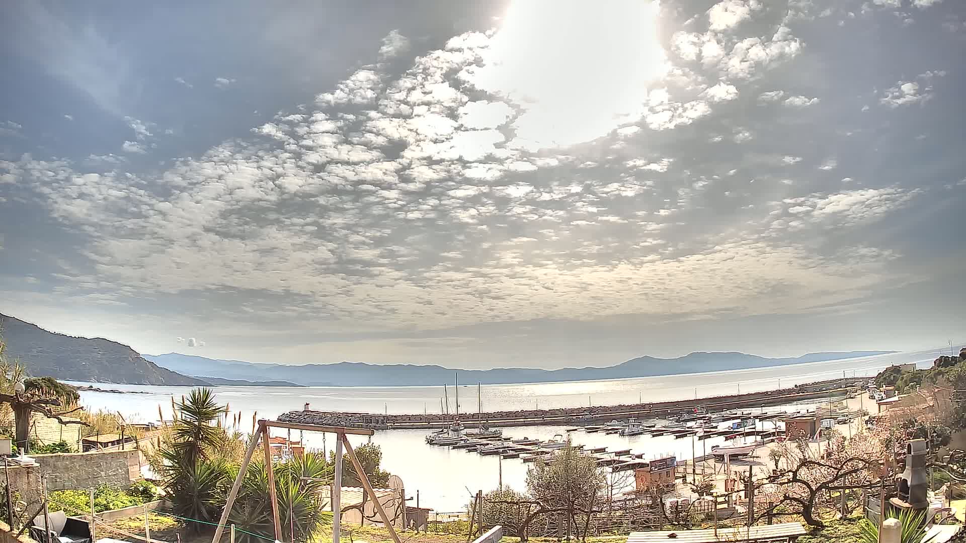 A partly sunny day overlooks a calm bay with a marina filled with boats, mountains in the distance, and a garden in the foreground.