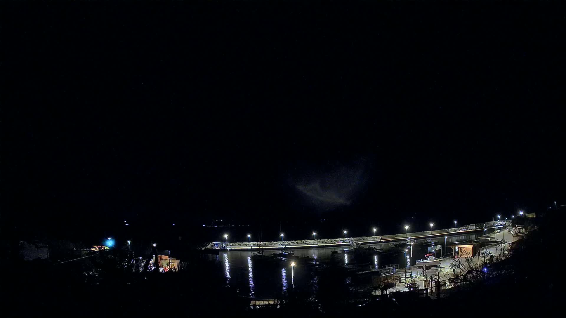 A nighttime view of a harbor with boats docked along a lit waterfront under a dark, mostly clear sky.