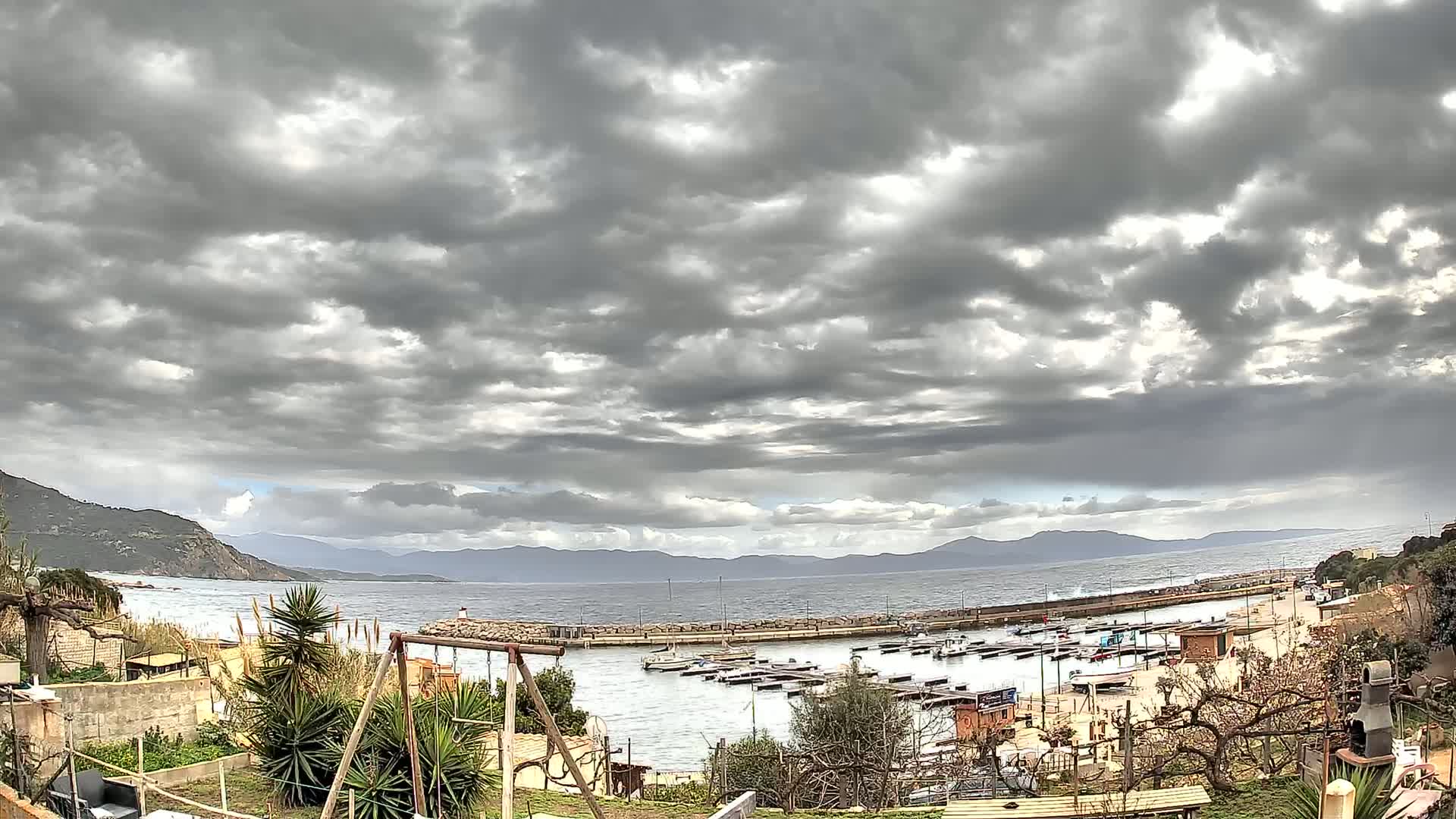 A small harbor with numerous boats is nestled between a rocky shore and distant mountains under a heavily clouded sky.