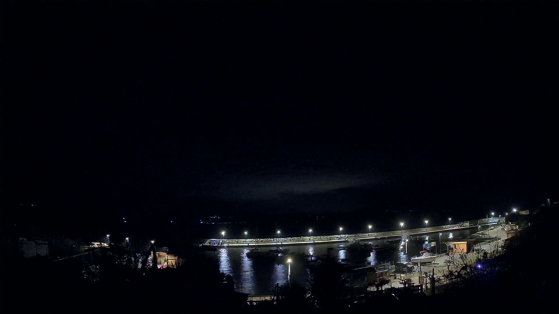 A nighttime view of a harbor with boats moored alongside a lit seawall under a dark, mostly clear sky.