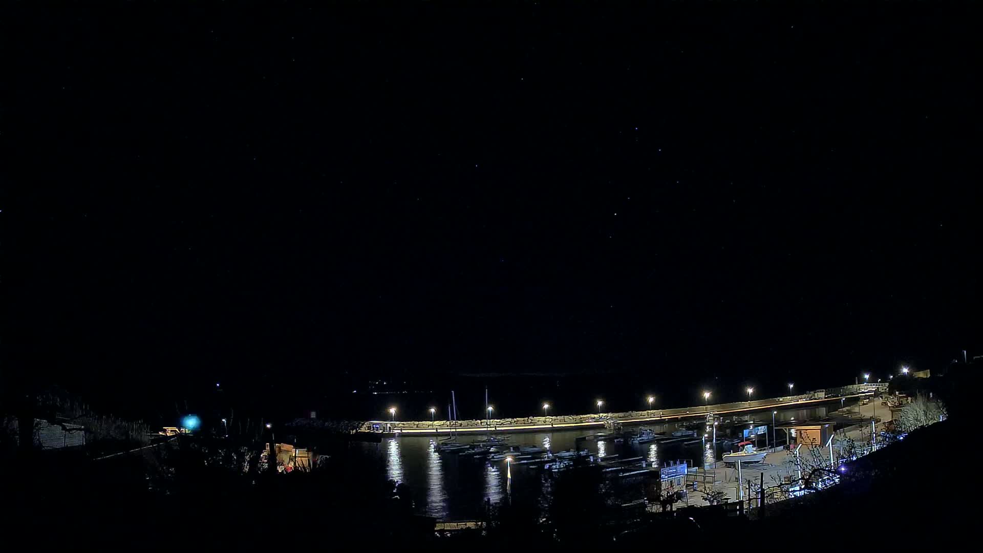 A nighttime view of a harbor filled with boats, illuminated by lights under a clear, starry sky.