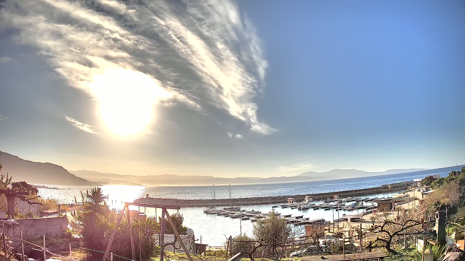 A sunny day with wispy clouds over a calm sea, featuring a small marina filled with boats, backed by hills and mountains in the distance.