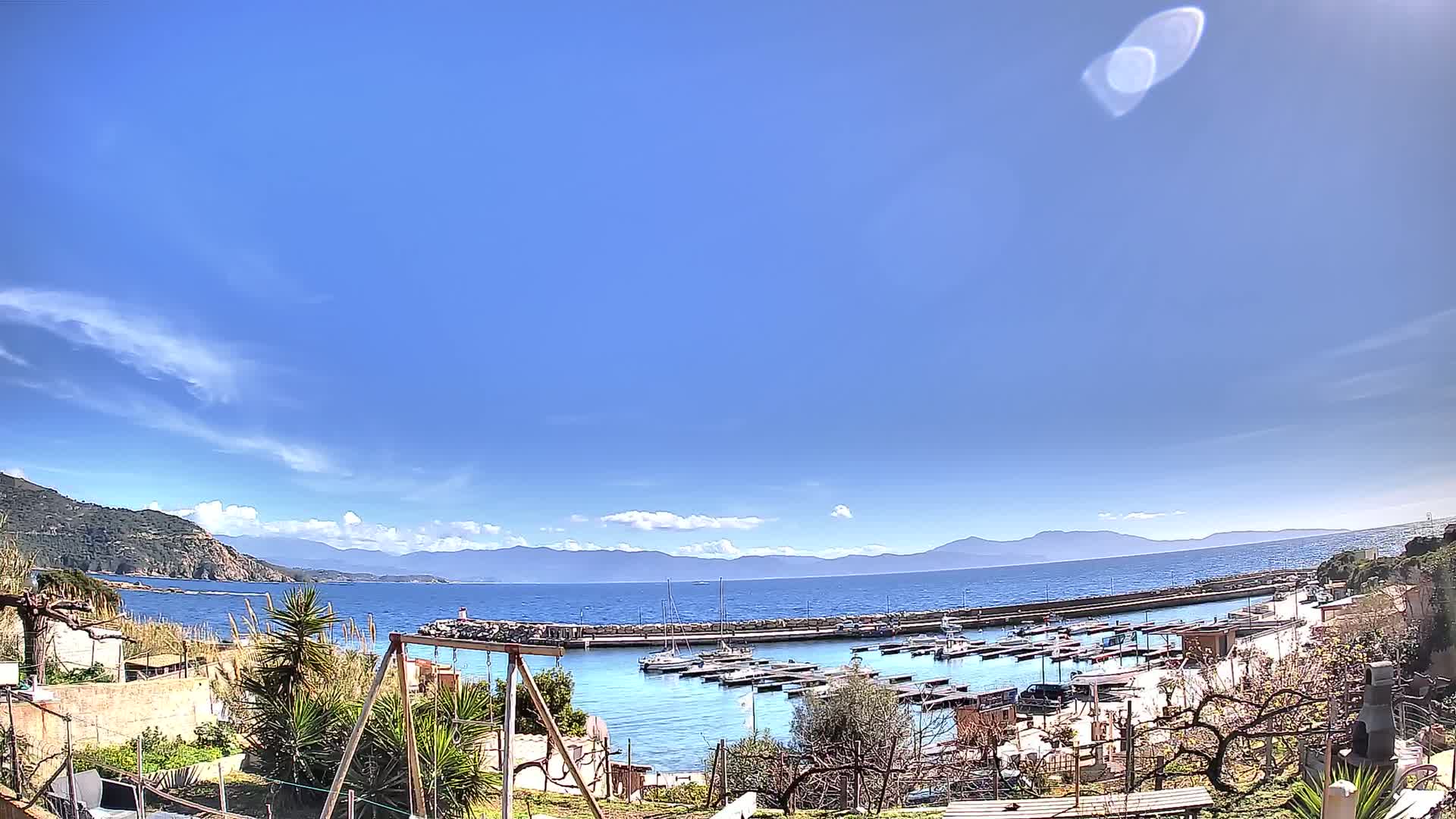 A small harbor filled with boats is seen from a hillside overlooking a calm sea under a mostly clear blue sky.