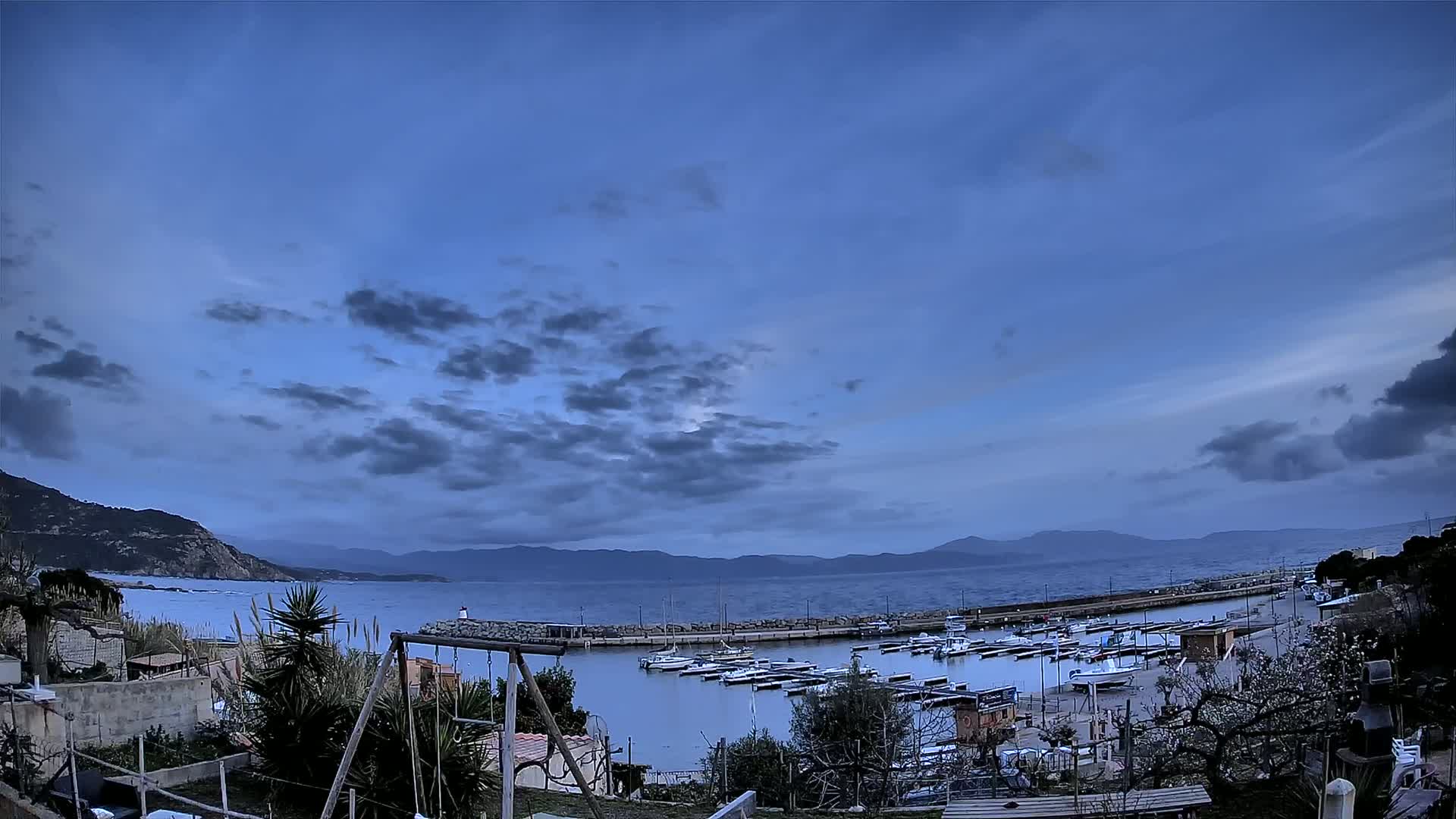 A small harbor filled with boats is nestled between a rocky hillside and a calm sea under a partly cloudy twilight sky.