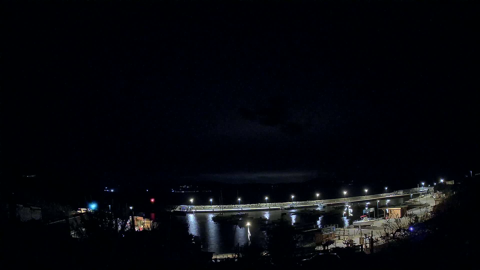 A nighttime view of a harbor with boats docked along a lit pier under a dark, cloudy sky.