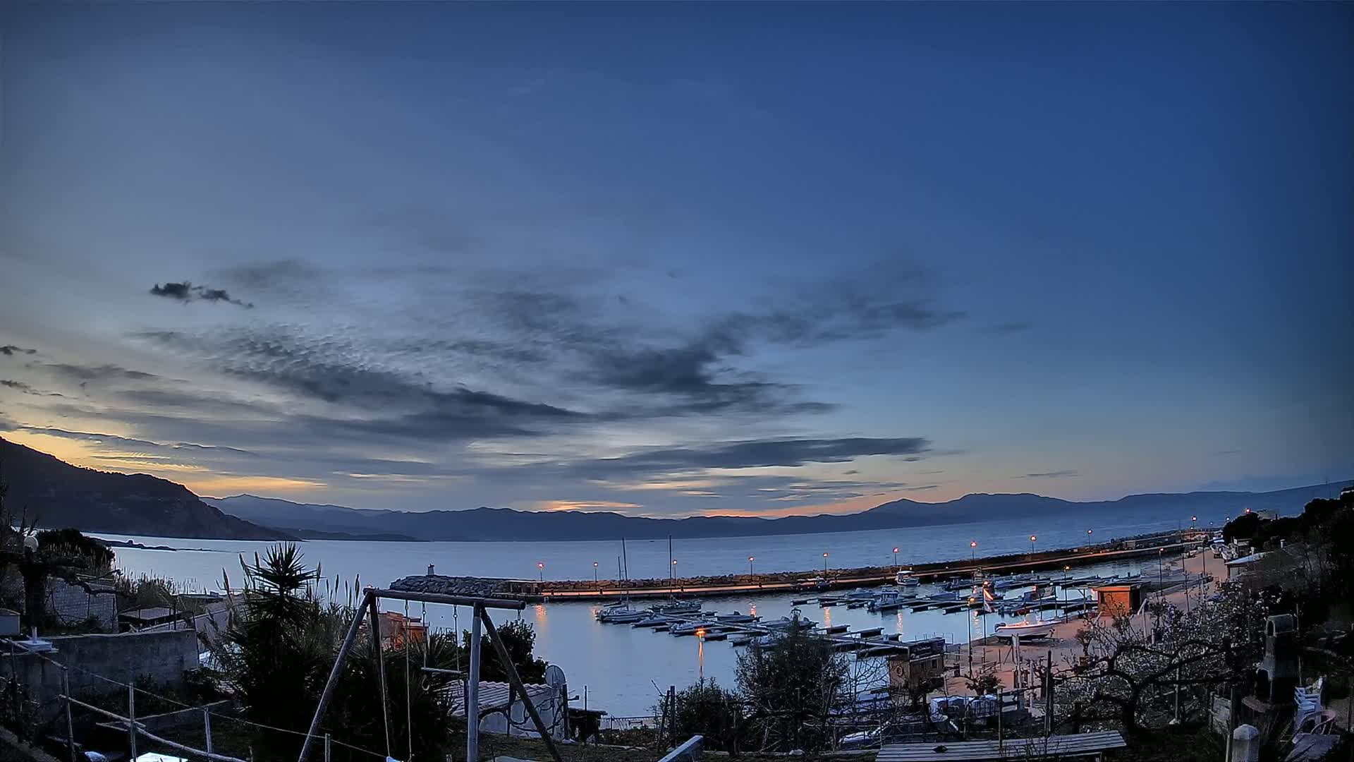 A twilight view of a calm harbor filled with boats, situated near a coastline with mountains in the background under a cloudy sky.