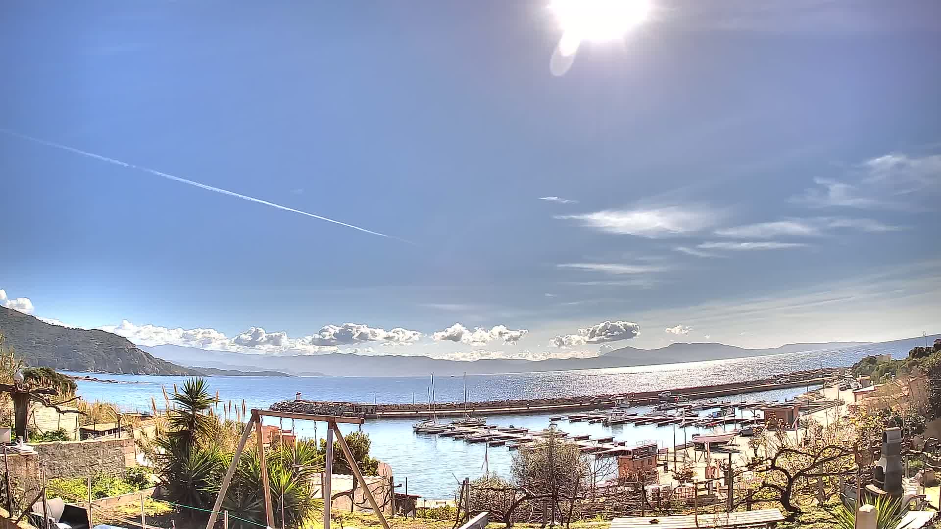 A sunny day with scattered clouds overlooks a calm bay filled with boats, nestled between a rocky coastline and distant mountains.